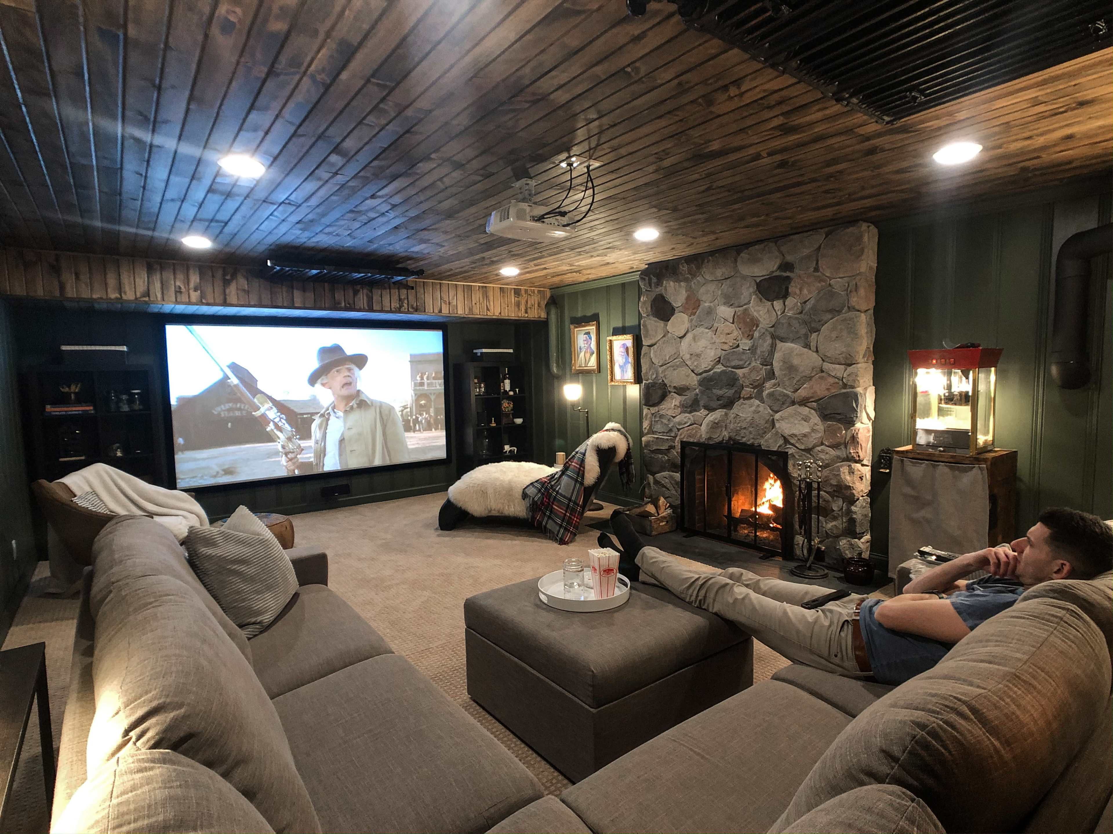 A person relaxes on a grey sectional sofa while watching a movie on a large screen in a cozy, stone-walled basement with a fireplace.