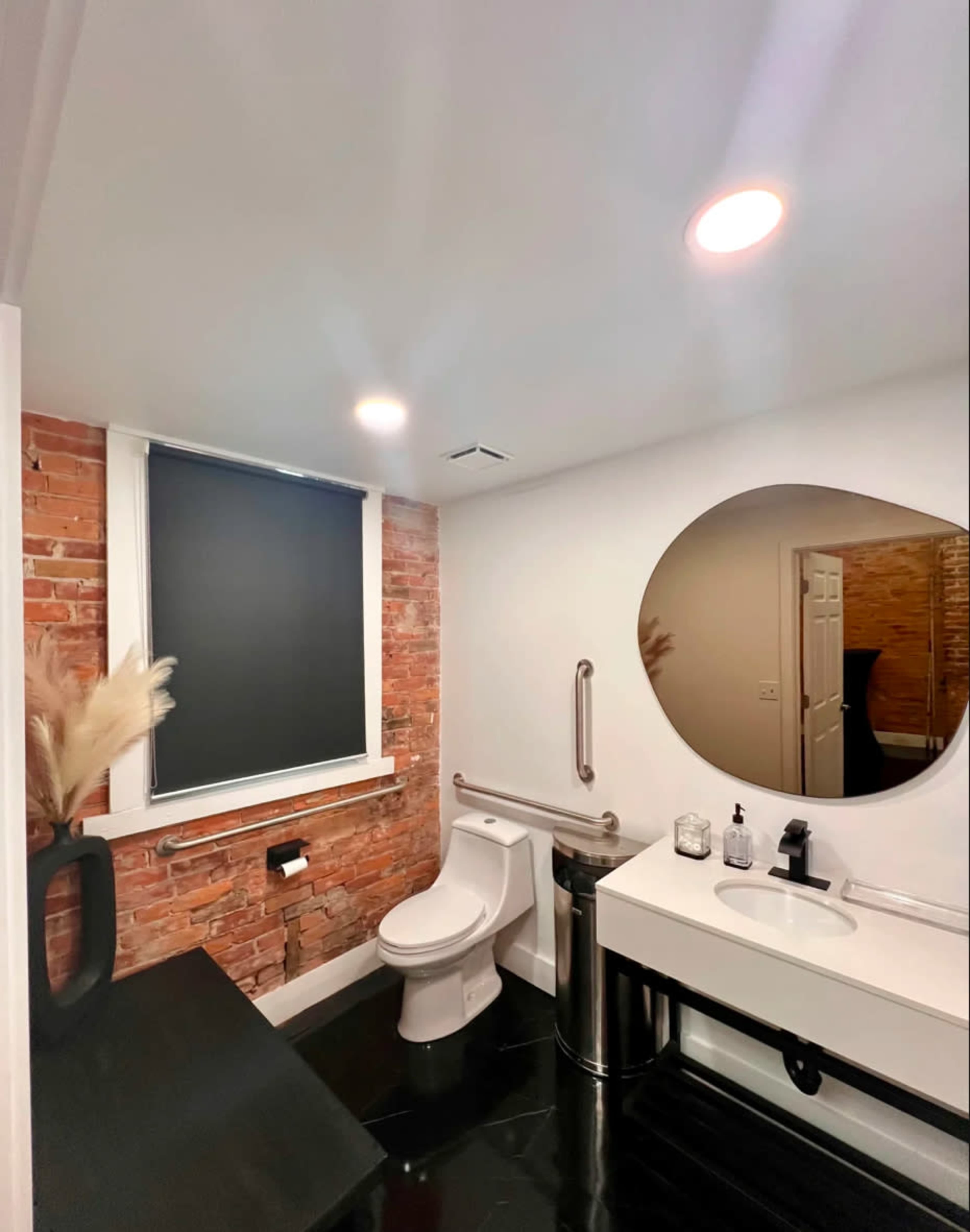 The bathroom features exposed brick walls, a circular mirror above a modern sink, and minimalistic fixtures.