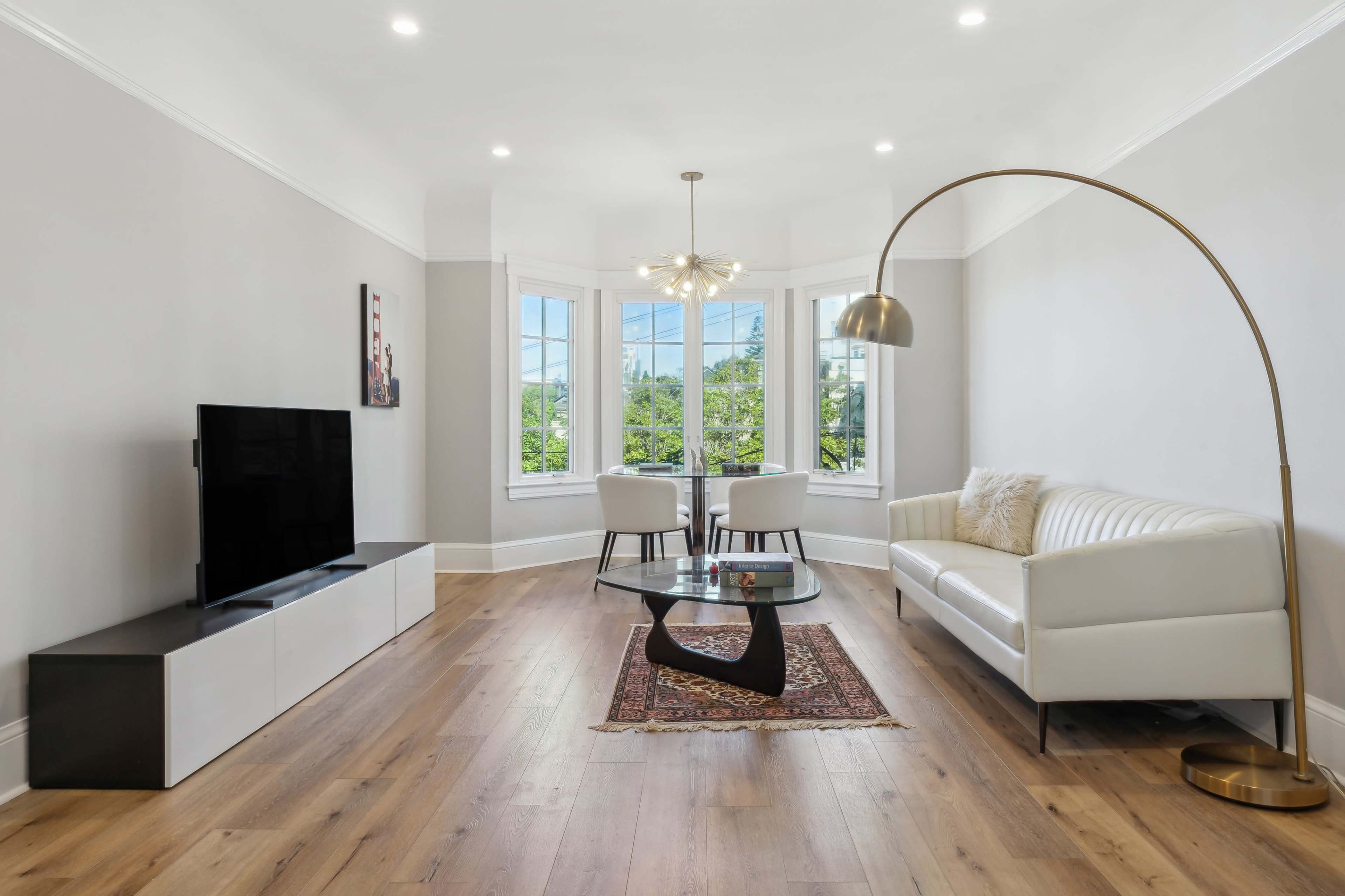 A modern living room features a white sofa, a glass coffee table, a television, and two chairs near large windows.