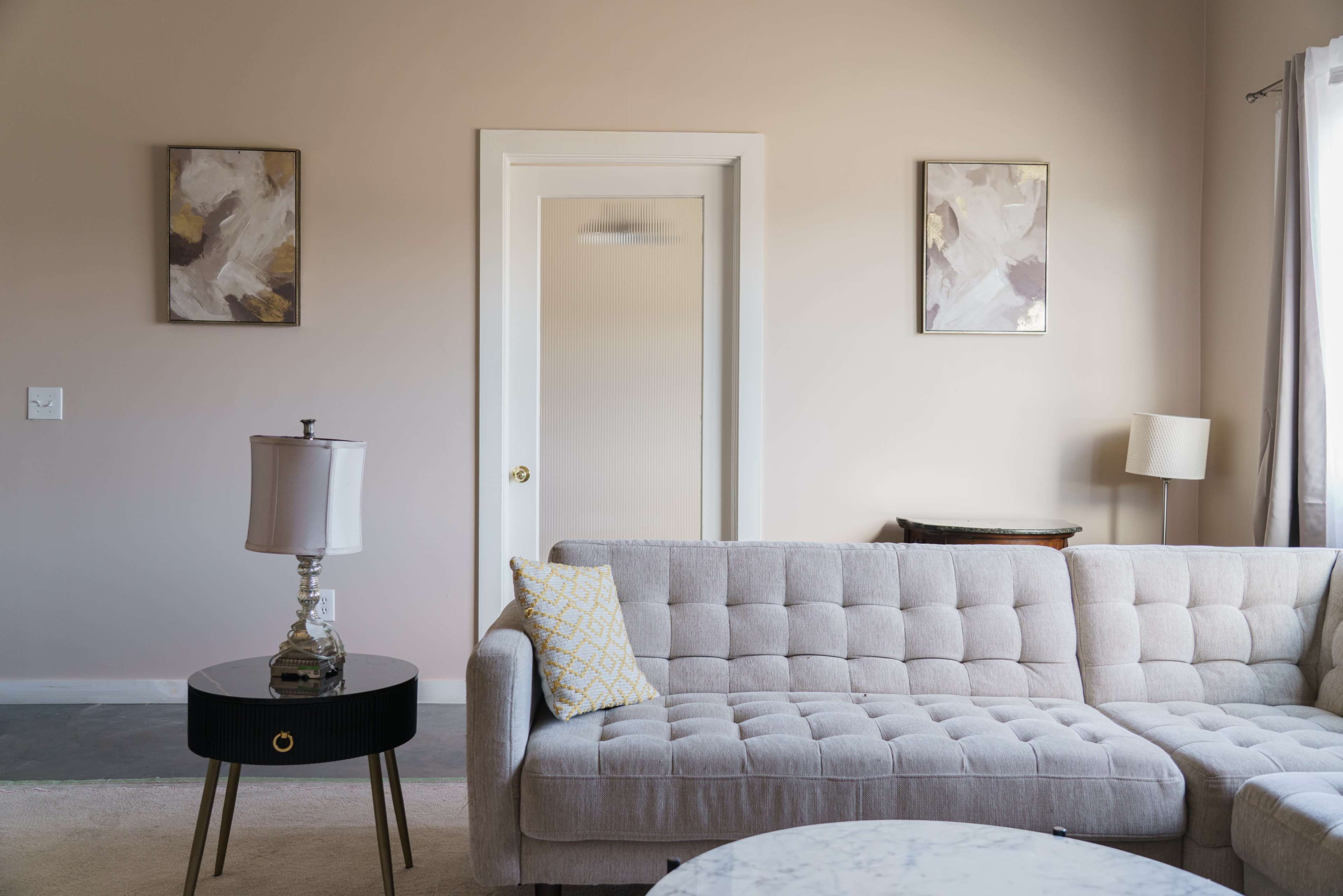 A light-colored living room features a gray tufted sofa, a small black side table with a lamp, and artwork hanging on the walls.