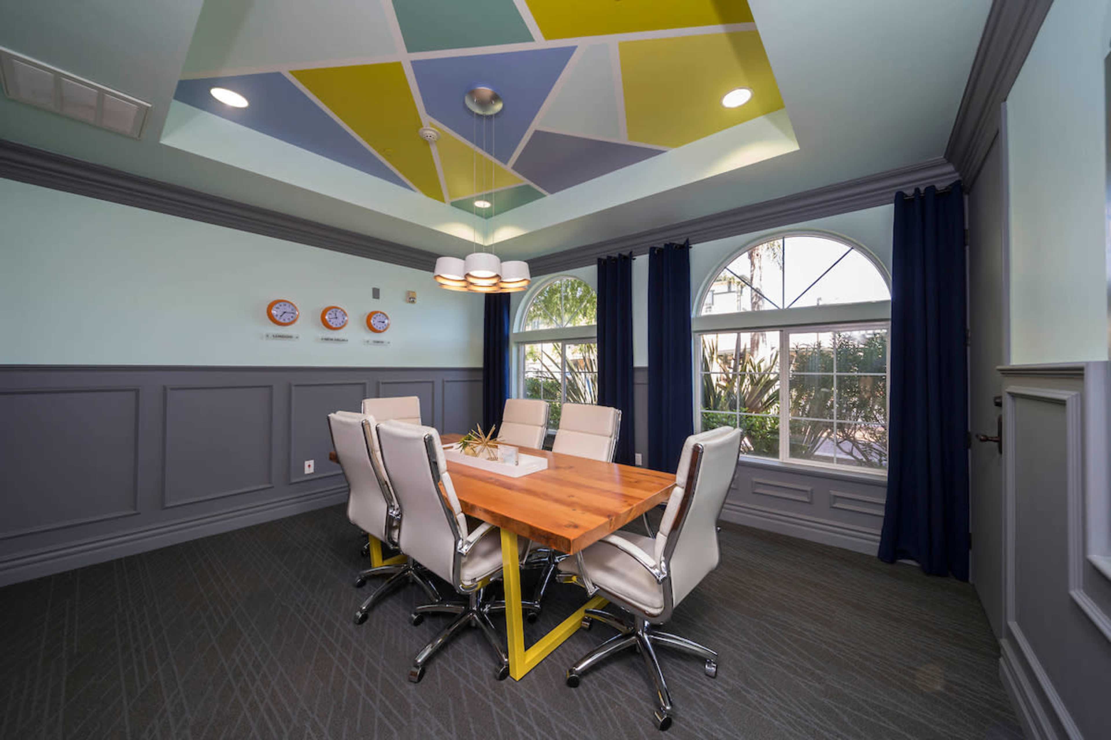 A conference room features a wooden table surrounded by white chairs, with a colorful ceiling and large windows overlooking greenery.