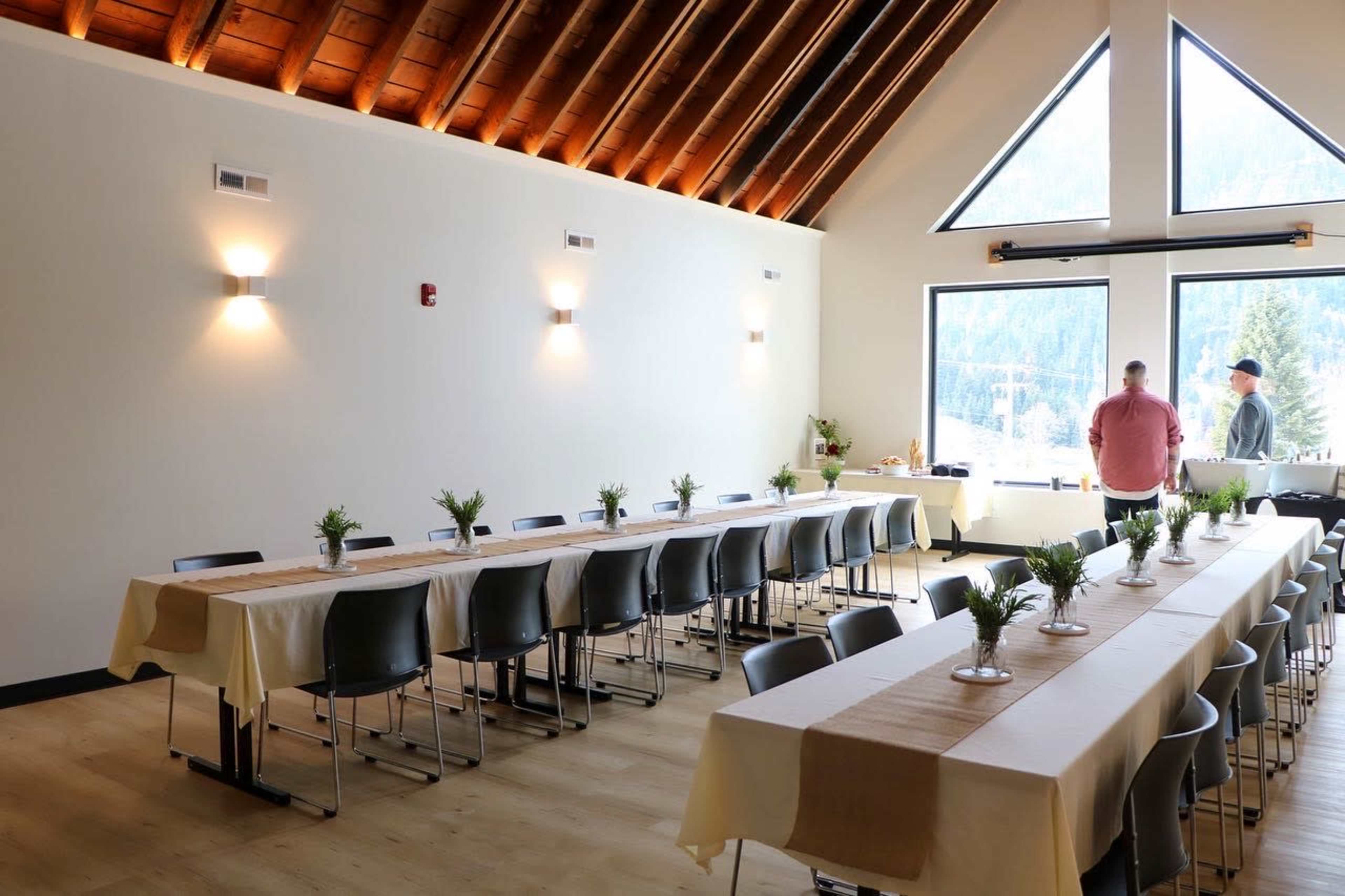 The image shows a spacious dining area with tables arranged in two long rows, each adorned with small plants, and large windows revealing a mountainous view.