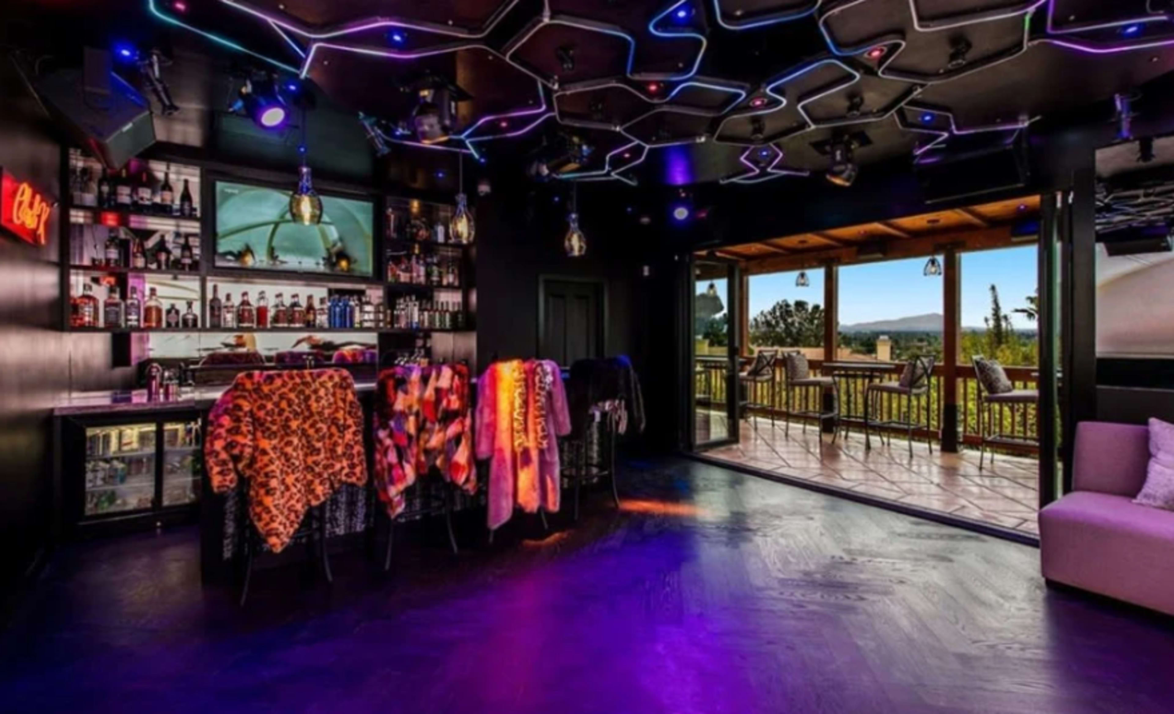 A modern bar area with colorful garments draped over bar stools, illuminated by vibrant lighting and featuring large glass doors that open to a scenic outdoor view.