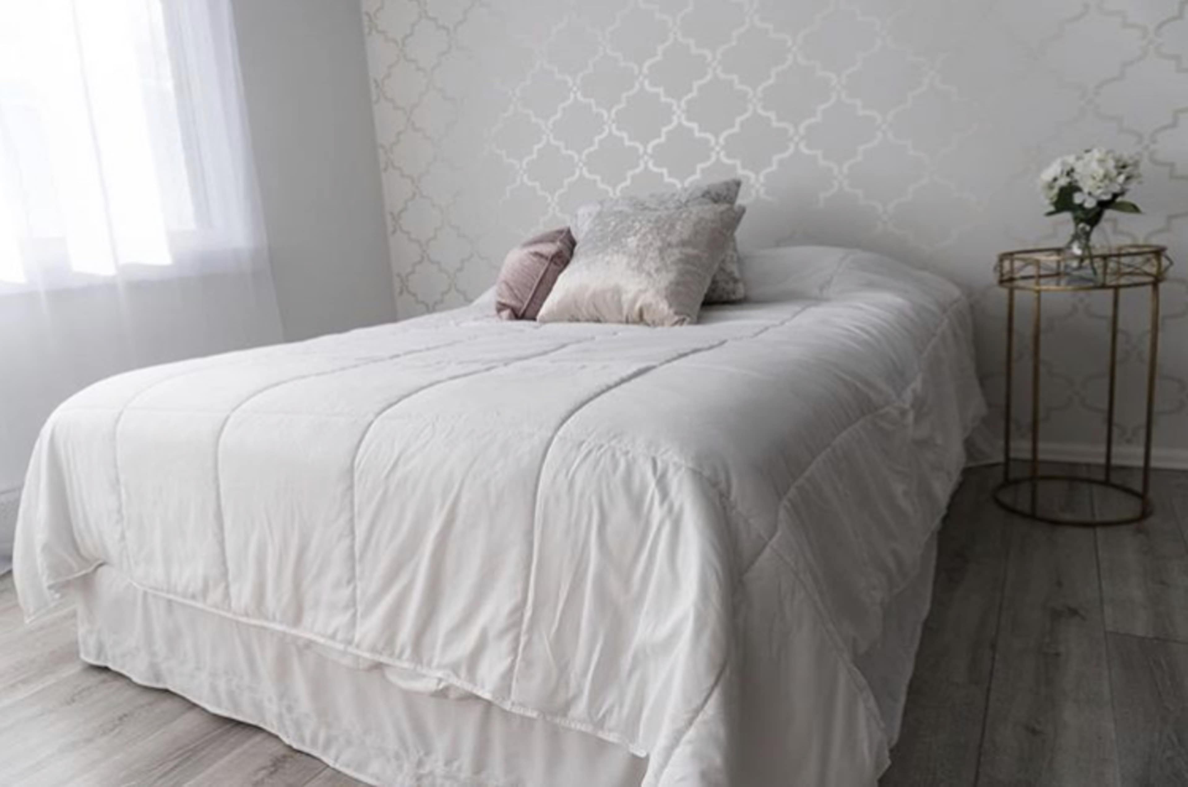 A neatly made bed with a white comforter, two decorative pillows, and a small side table with a vase of flowers in a well-lit room.