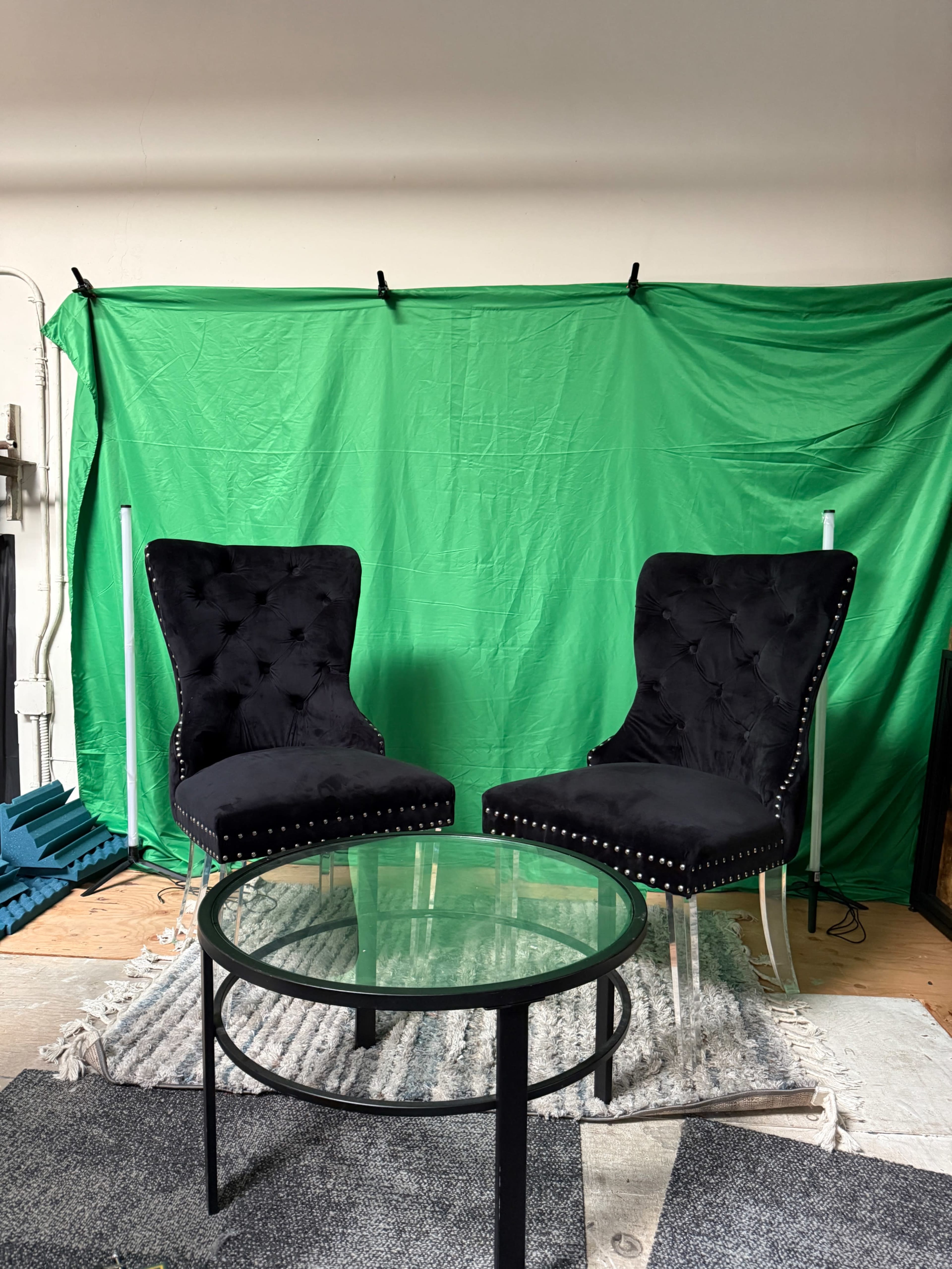 Two black velvet chairs with tufted backs are positioned on either side of a glass table in front of a green backdrop.