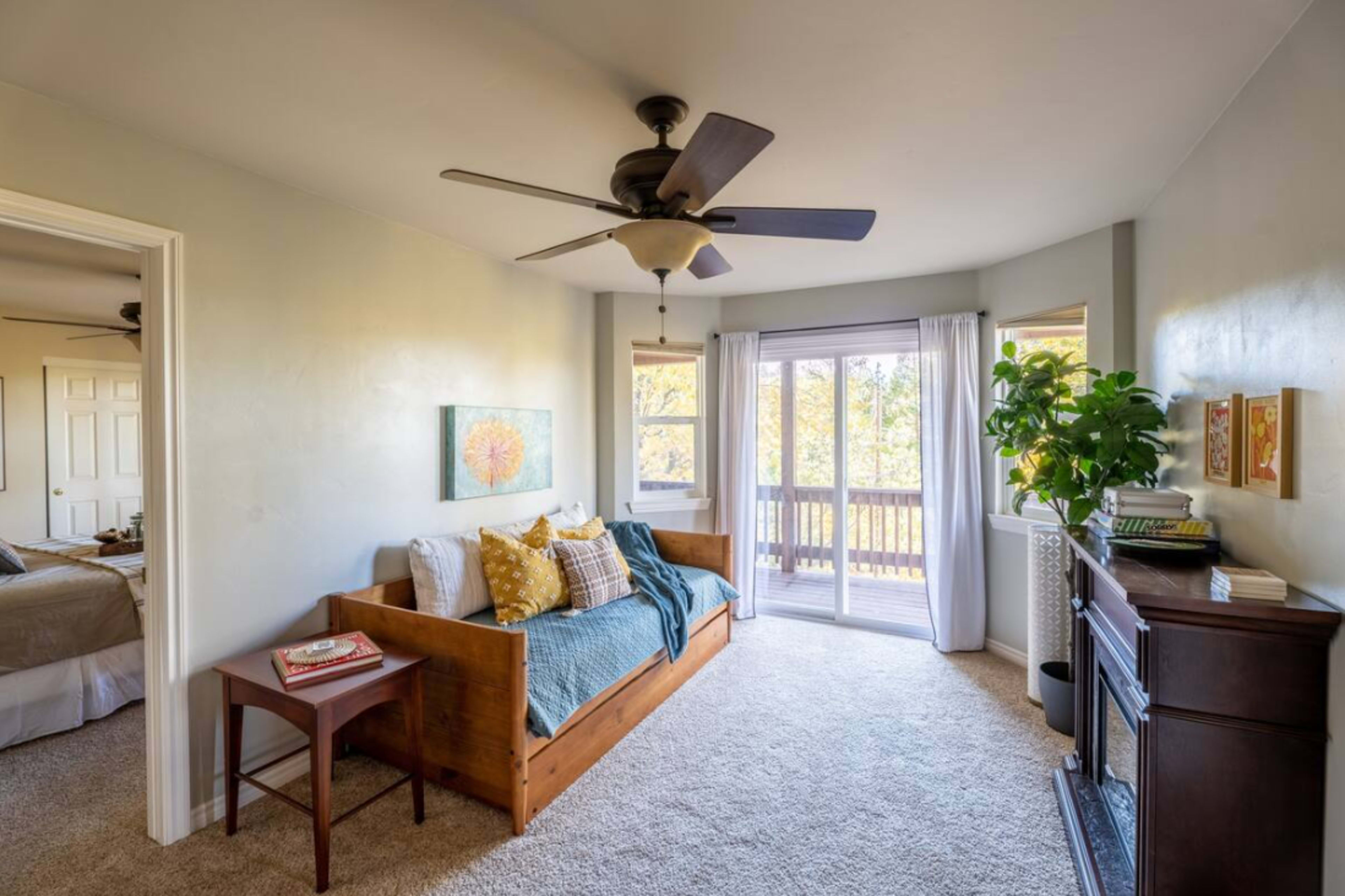A well-lit bedroom featuring a daybed with decorative pillows, a ceiling fan, a small side table, and a window with sheer curtains overlooking a balcony.