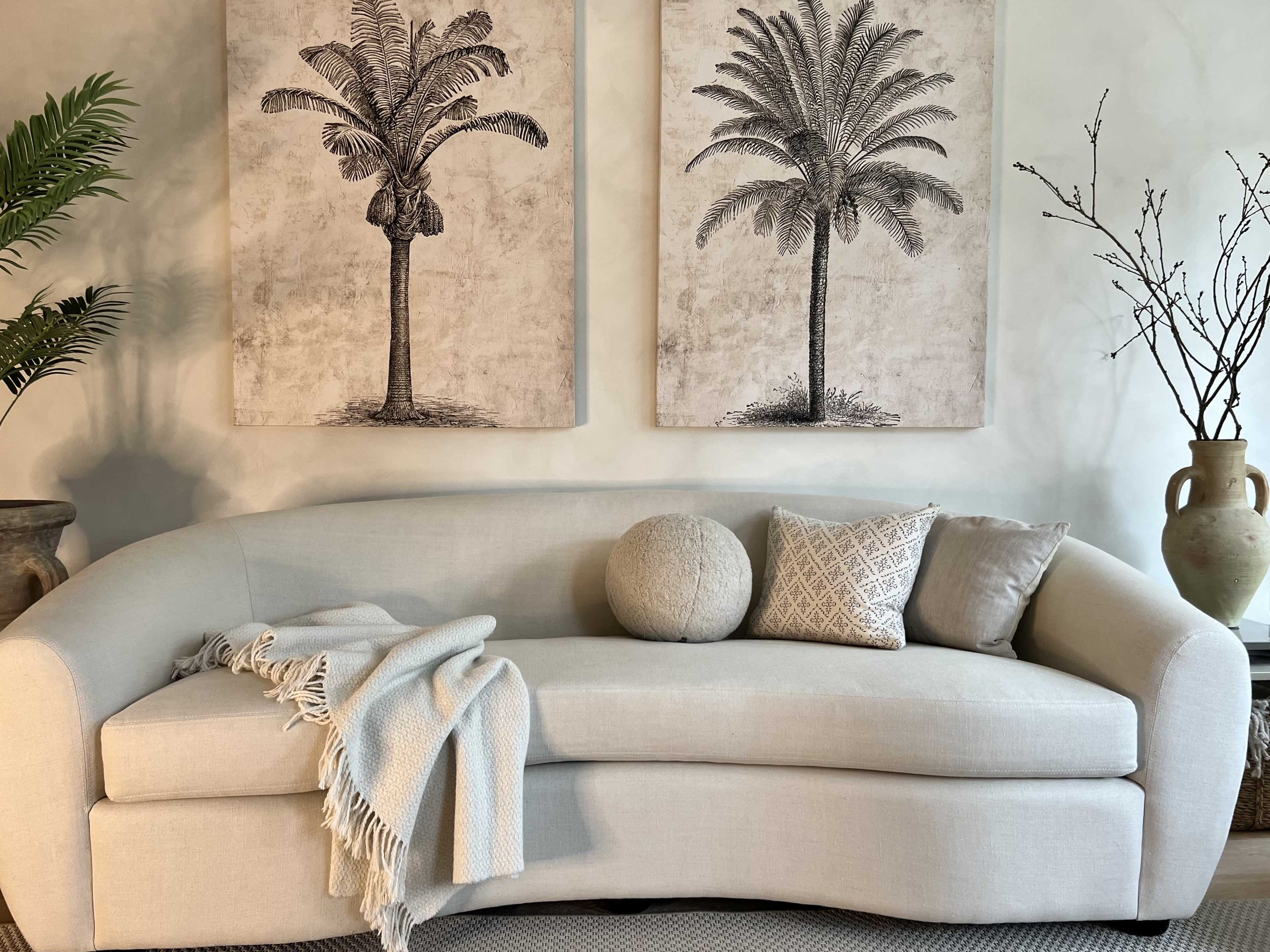 A light-colored curved sofa adorned with pillows and a throw blanket, positioned between two palm tree artworks against a neutral wall.