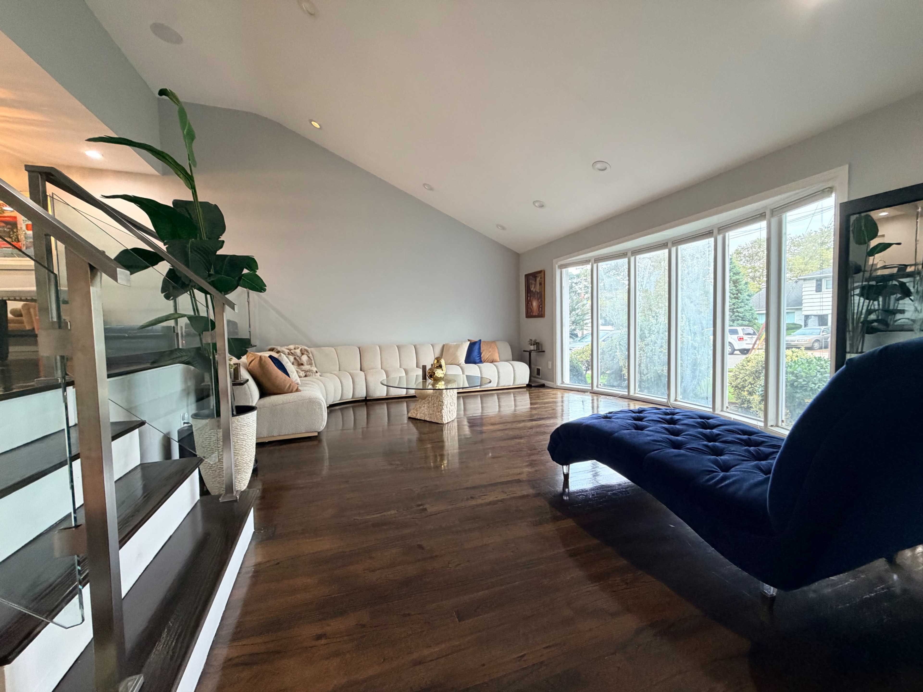 The living room features a light-colored sectional sofa, a blue lounge chair, a large bay window, and polished wooden flooring.