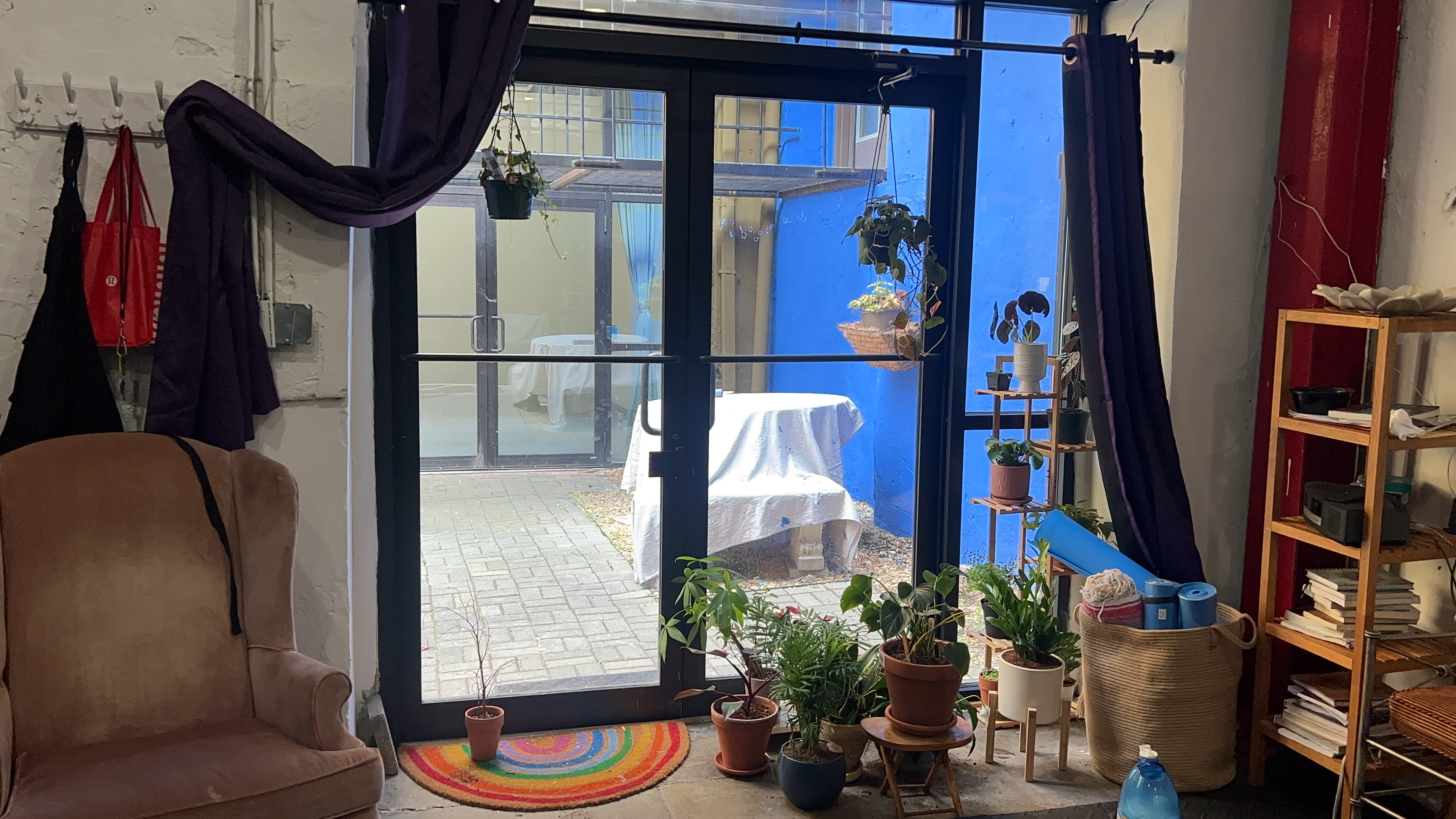 A cozy interior features a chair near a glass door with plants and a colorful rug, leading to a blue-walled outdoor space.