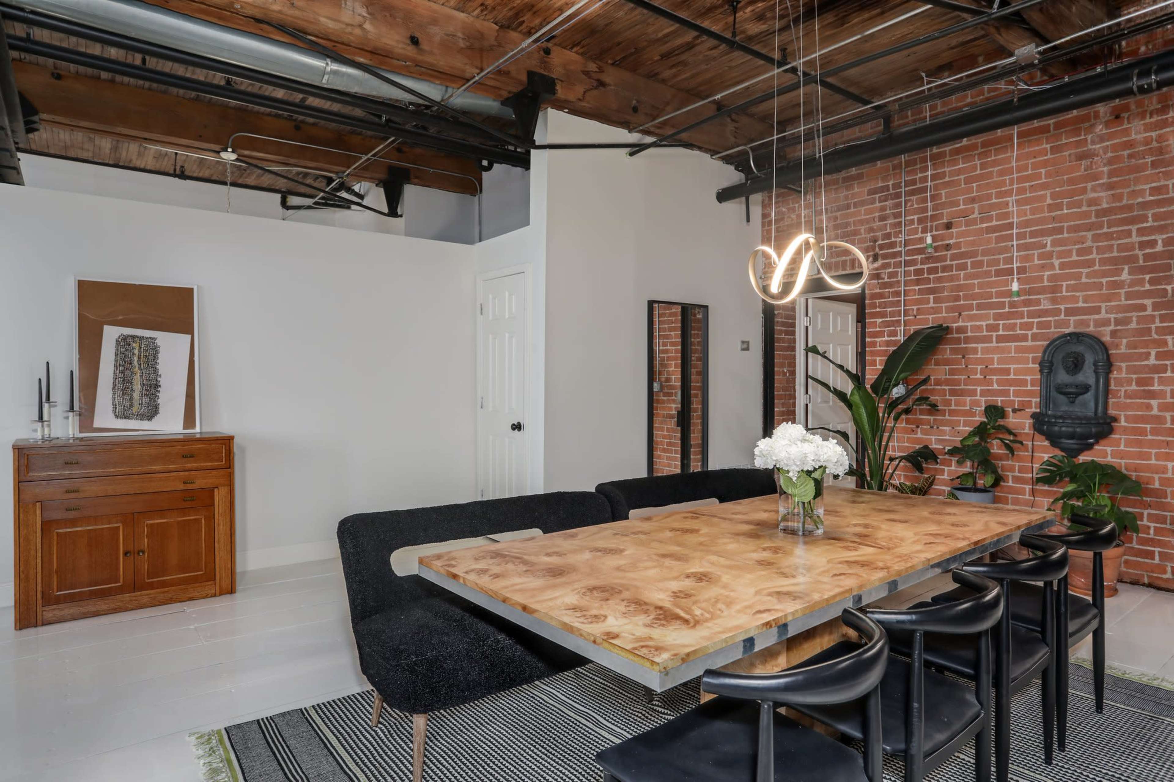 A modern dining area featuring a large wooden table surrounded by black chairs, with a brick wall in the background and a cabinet on the side.