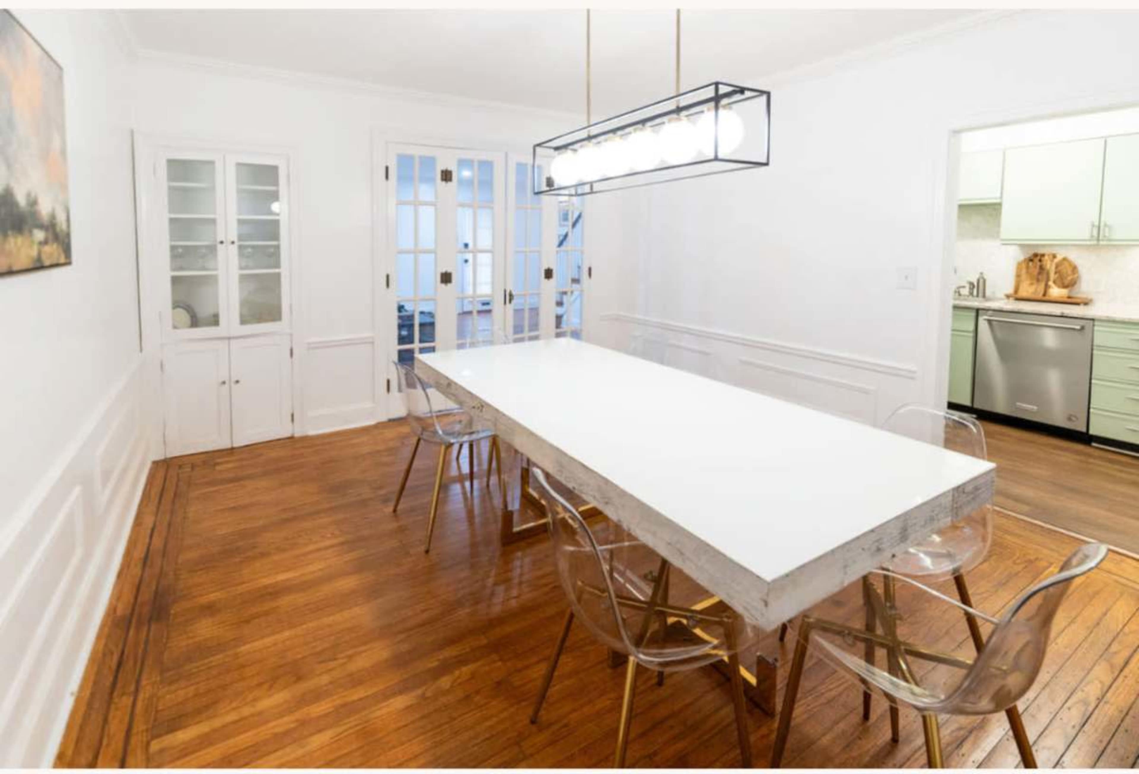 The image shows a modern dining room featuring a long white table surrounded by clear acrylic chairs, with a light fixture overhead and an open door leading to another room.