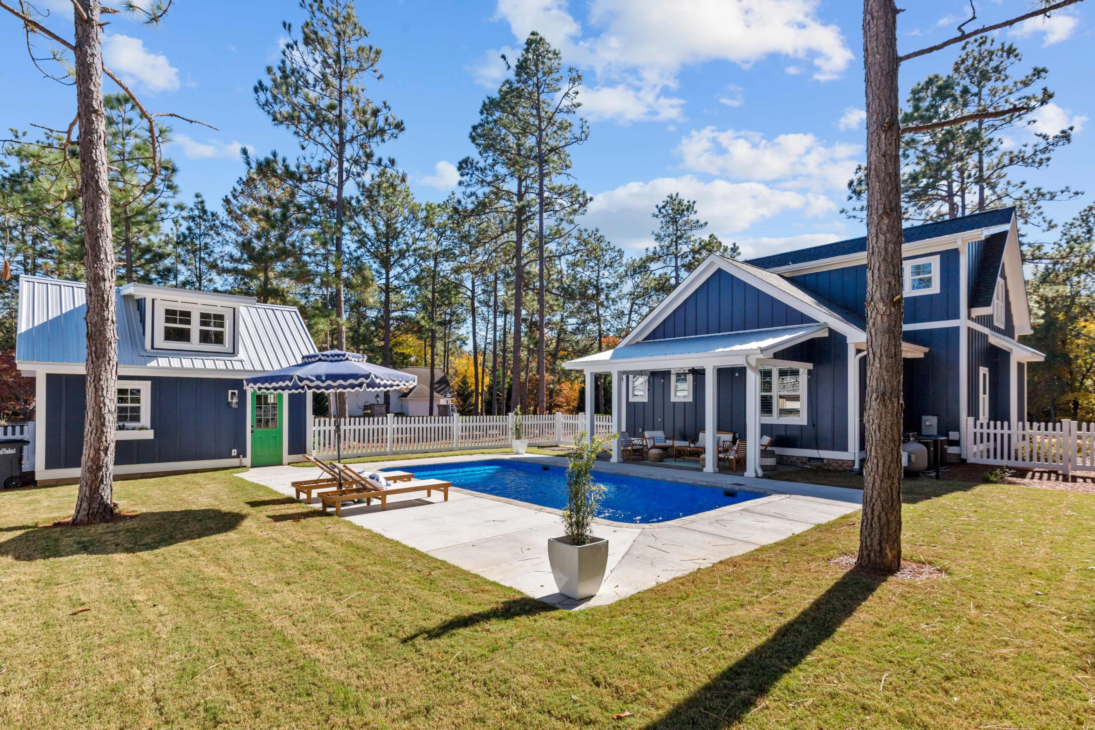 The image shows a modern house with a swimming pool surrounded by grass and tall trees, featuring a small guest house nearby.
