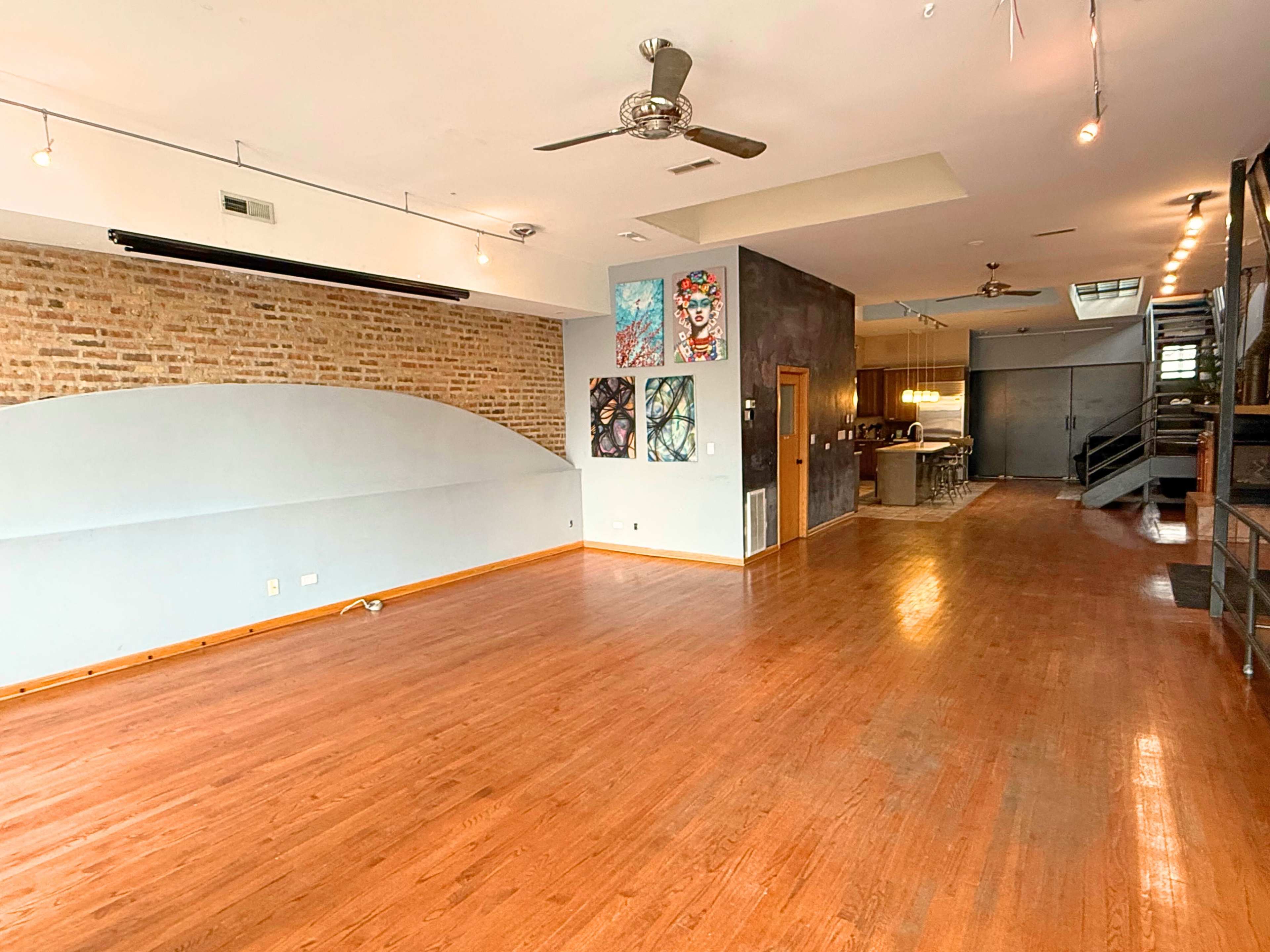 The image shows a spacious, modern interior with hardwood floors, exposed brick walls, and artwork displayed on one side, leading to a kitchen area in the background.