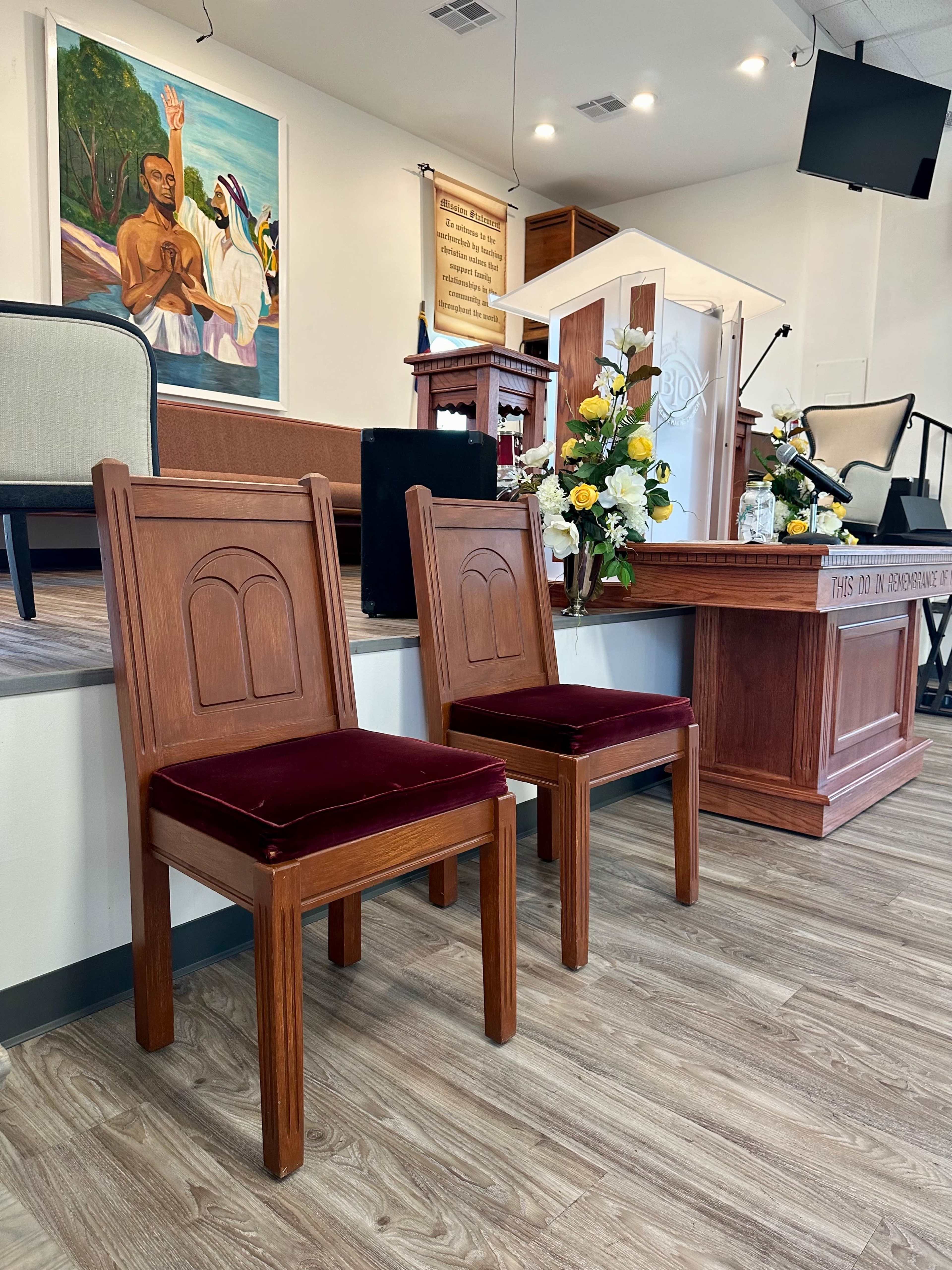 The image shows two wooden chairs with burgundy upholstery situated in a church setting, facing a podium and decorated with flowers.