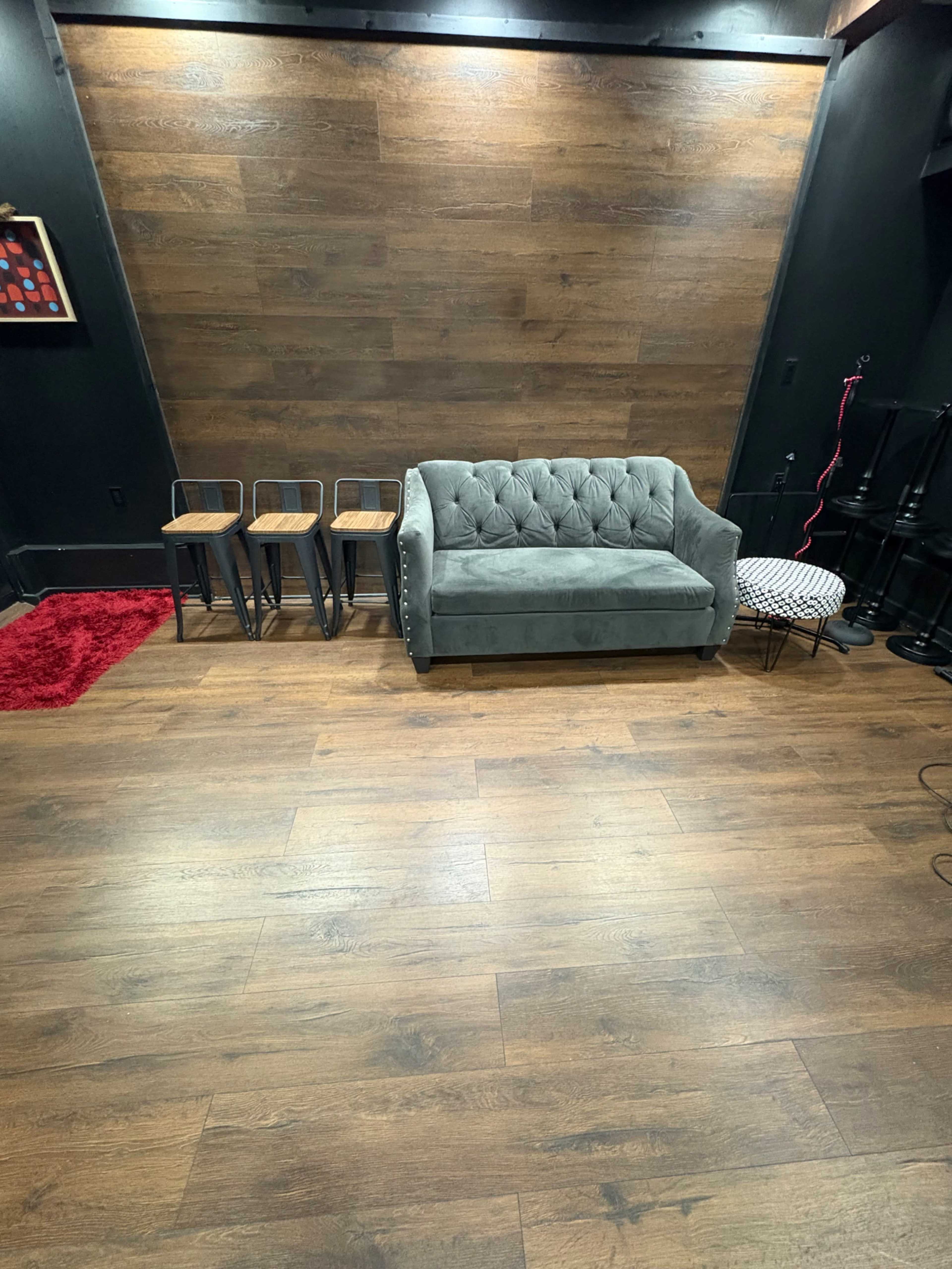 The image shows a modern interior space featuring a tufted gray sofa, three metal stools, and a small round table on a wooden floor with a dark wall behind.