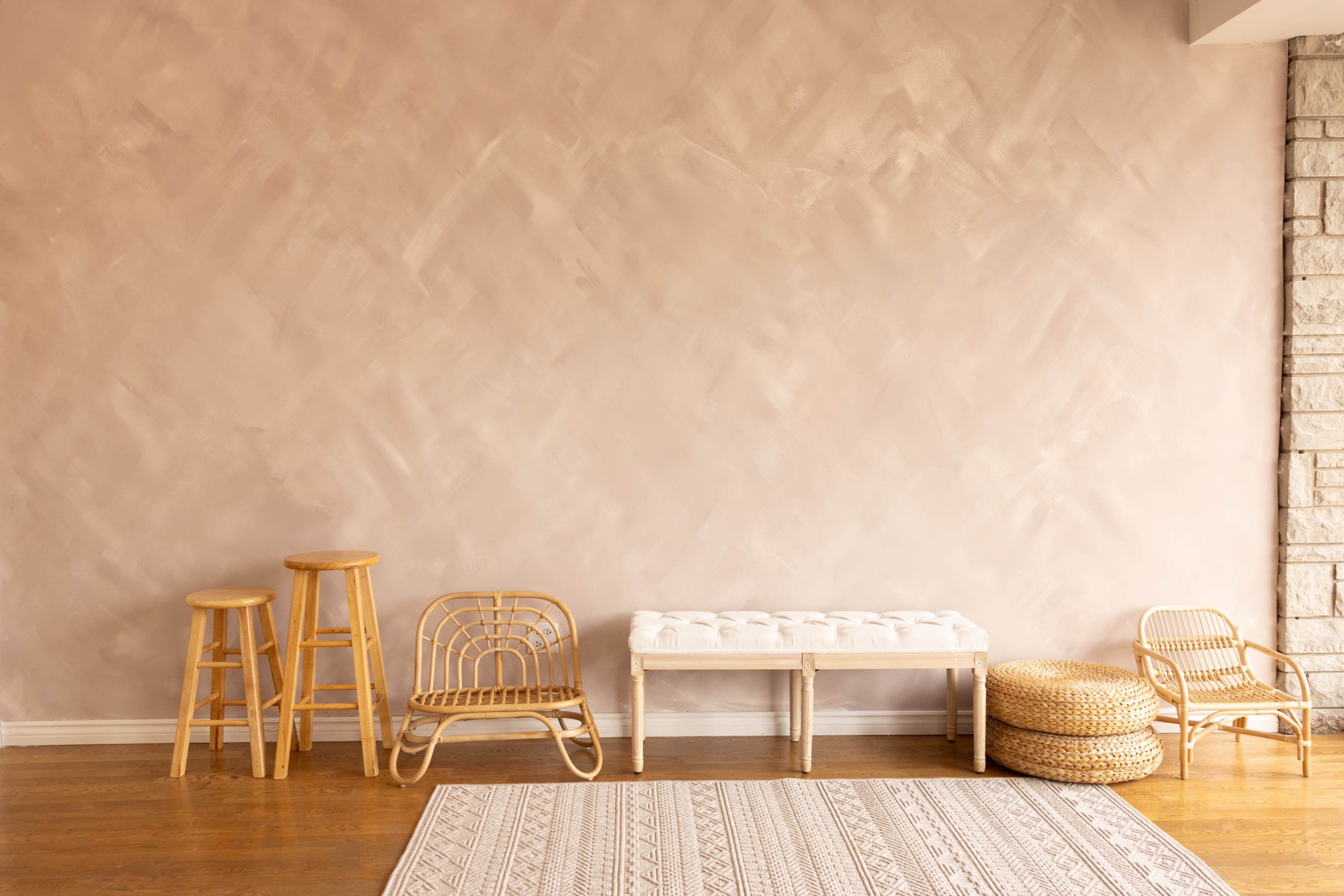 The image shows a minimalist interior with a textured pink wall, wooden flooring, a white tufted bench, and several pieces of rattan furniture.