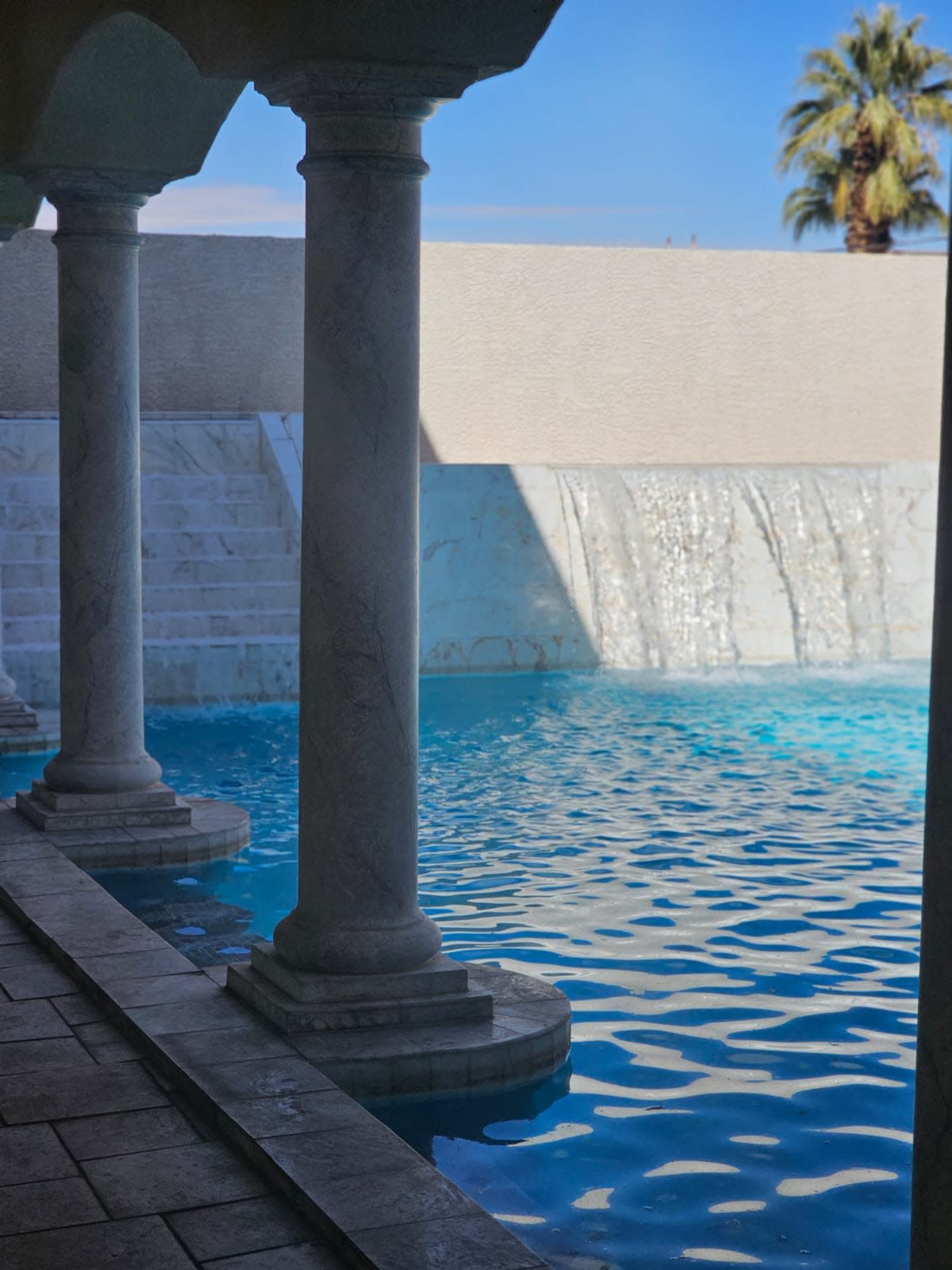 A pool area with marble columns and a waterfall, surrounded by water reflecting light under a blue sky.