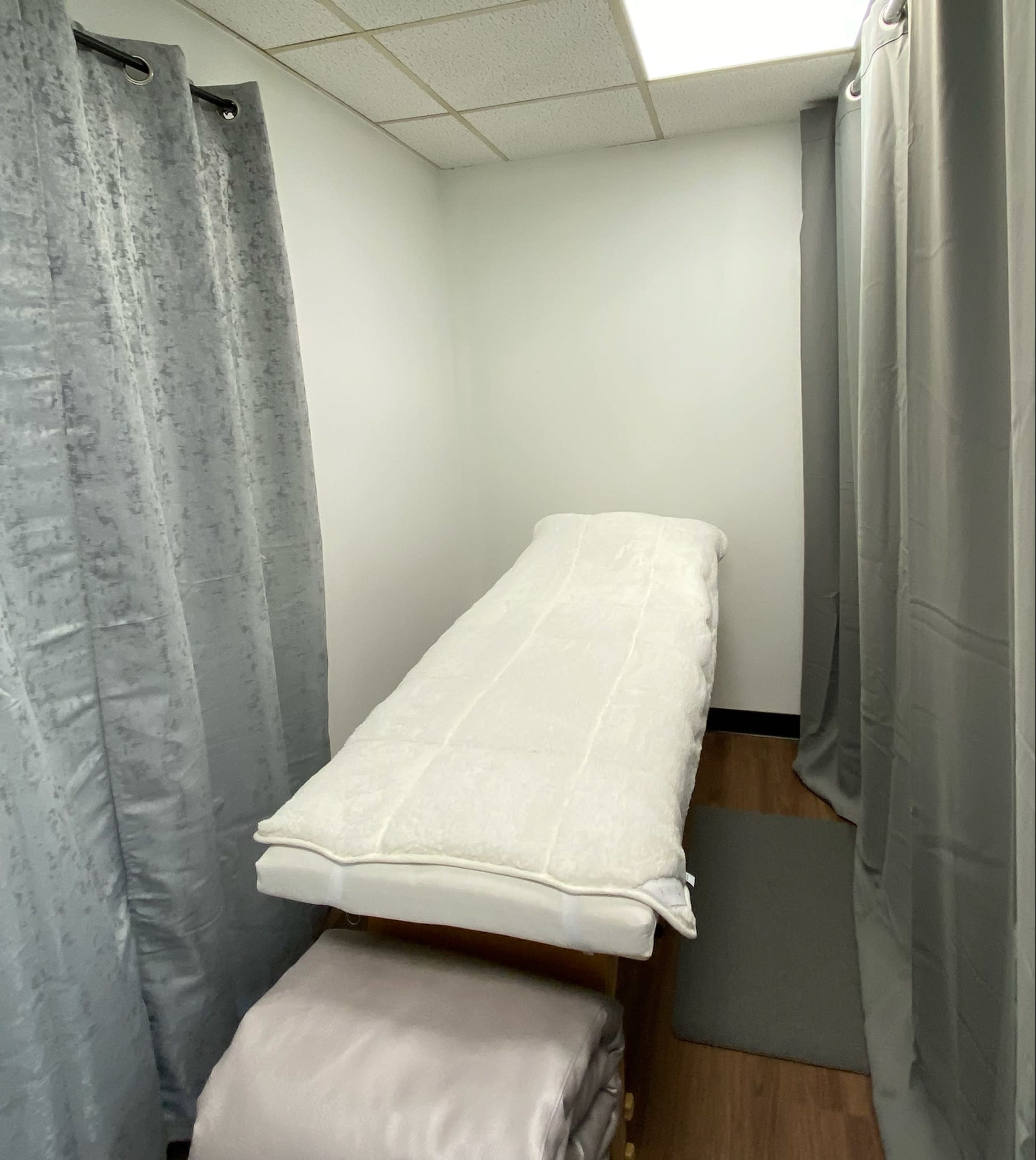 The image shows a small treatment room with a massage table covered in a white sheet, flanked by gray curtains.