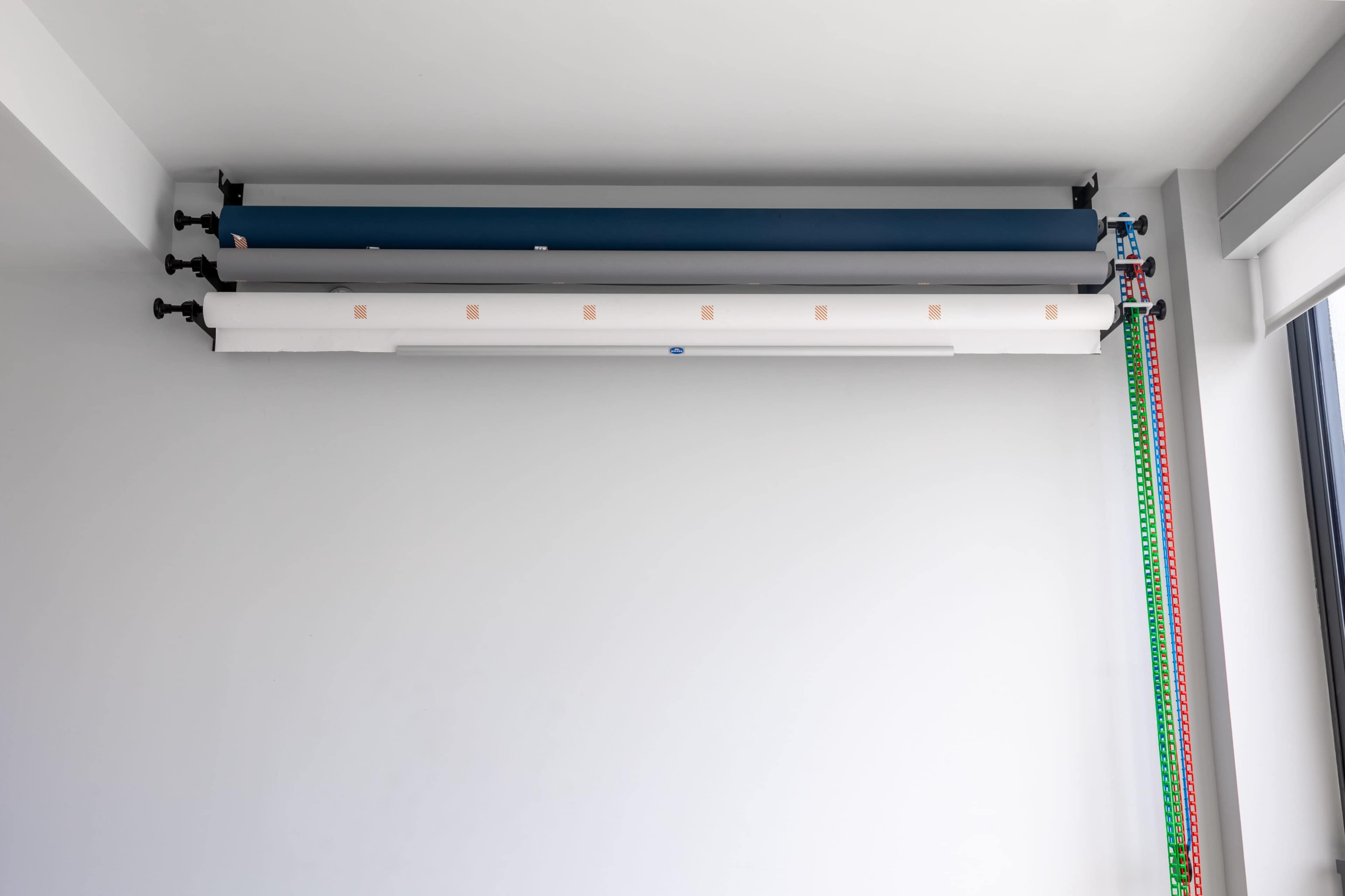 Three rolls of backdrop paper are stored horizontally along a wall, with one roll secured on a wall-mounted rack and a lighting strip visible on the right side.