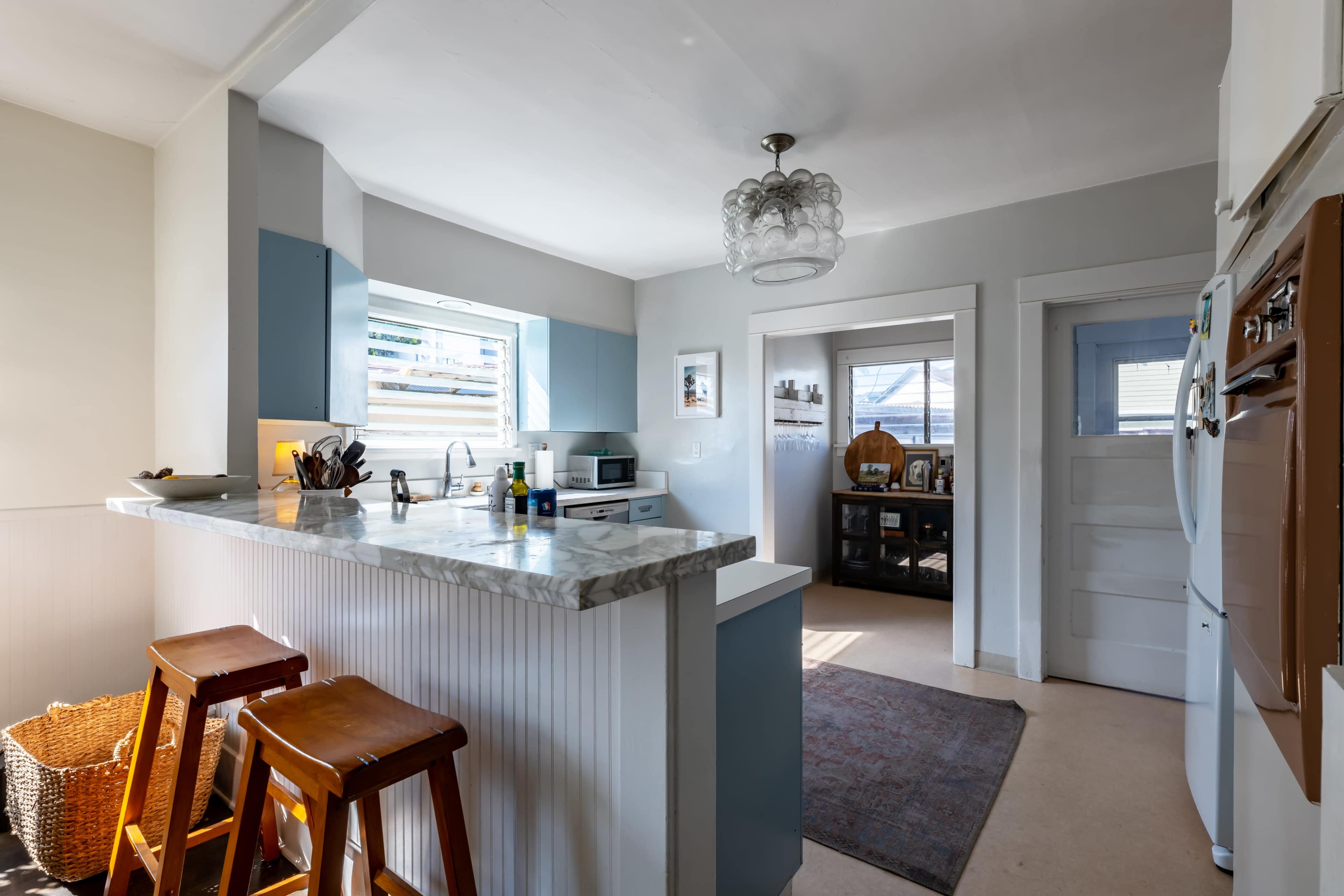A modern kitchen features a marble countertop island with wooden stools, open to a dining area through a doorway.