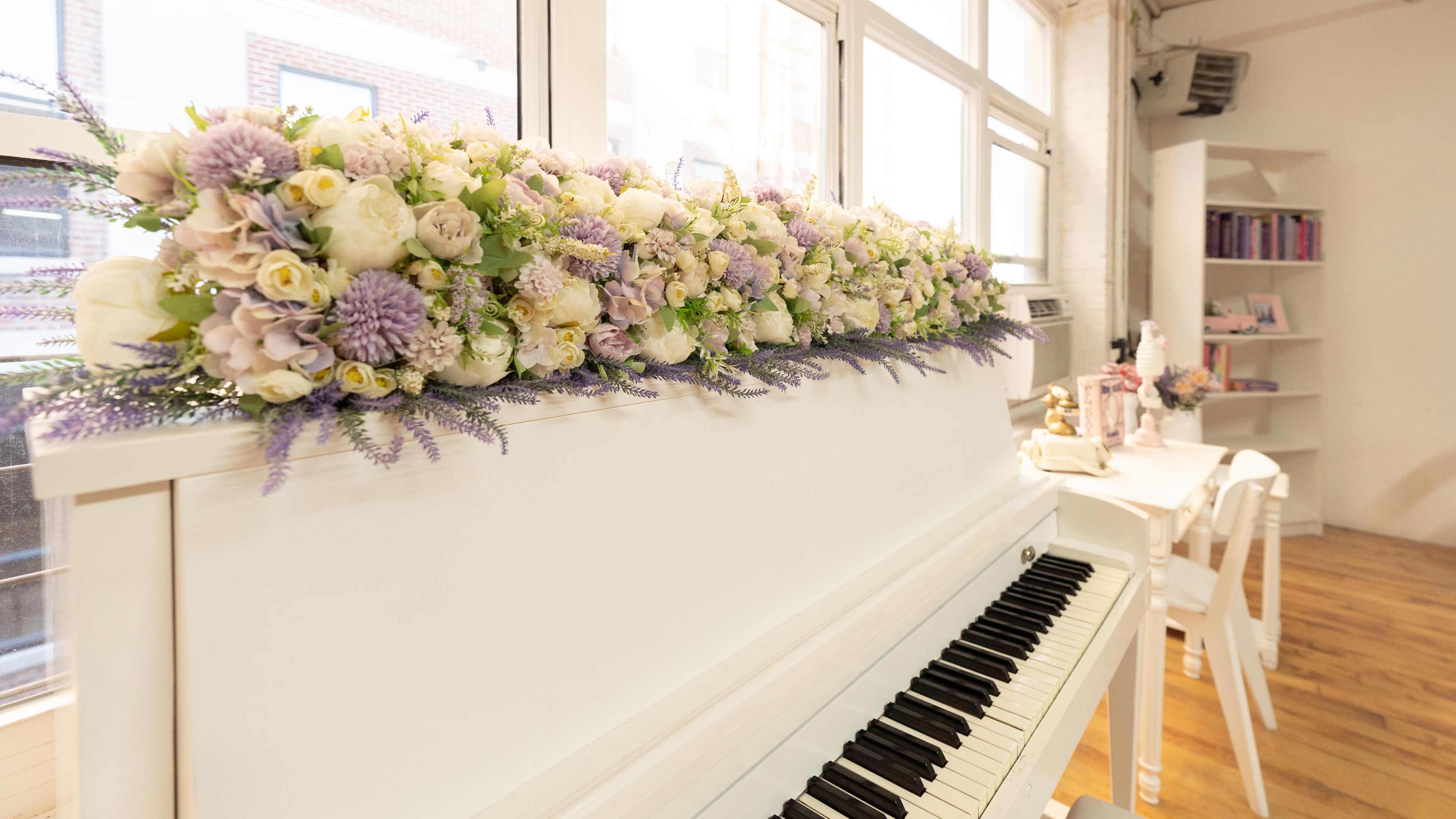 A white piano is adorned with a long arrangement of pastel flowers, while a light-colored table with decorative items is seen in the background.