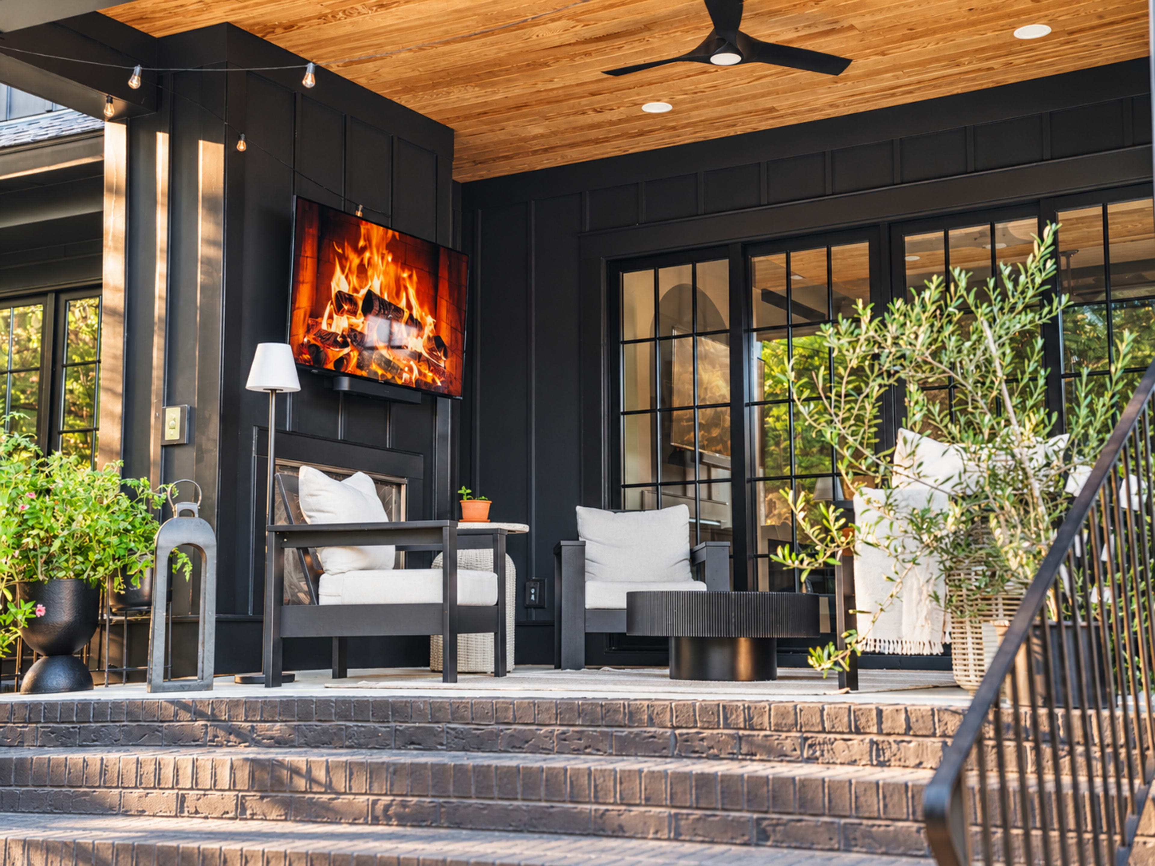 The image shows a modern porch with comfortable seating arrangements, a wall-mounted fireplace display, and greenery surrounding the area.