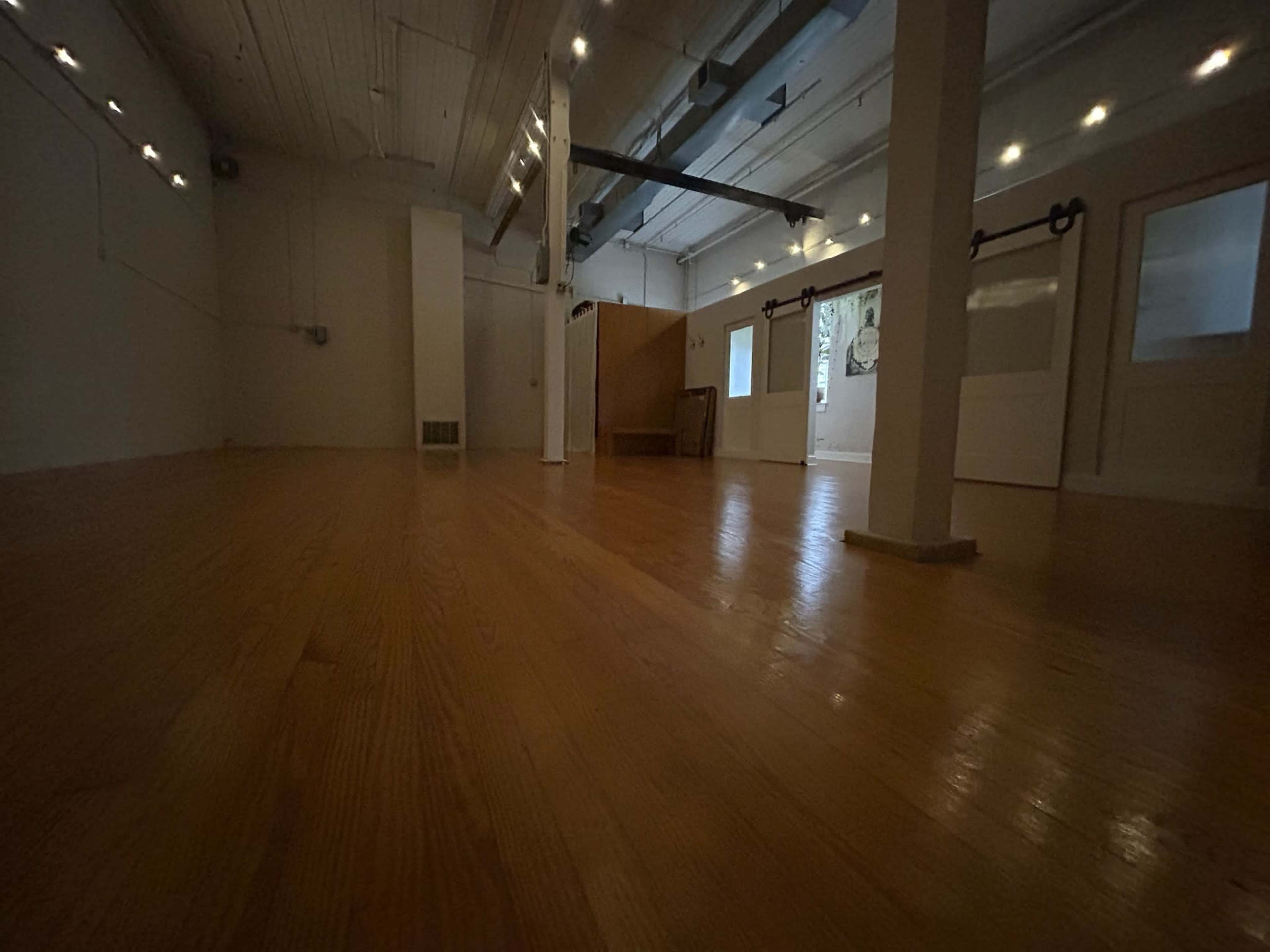 The image shows a spacious, empty room with wooden flooring and minimal furnishings, featuring natural light from windows.