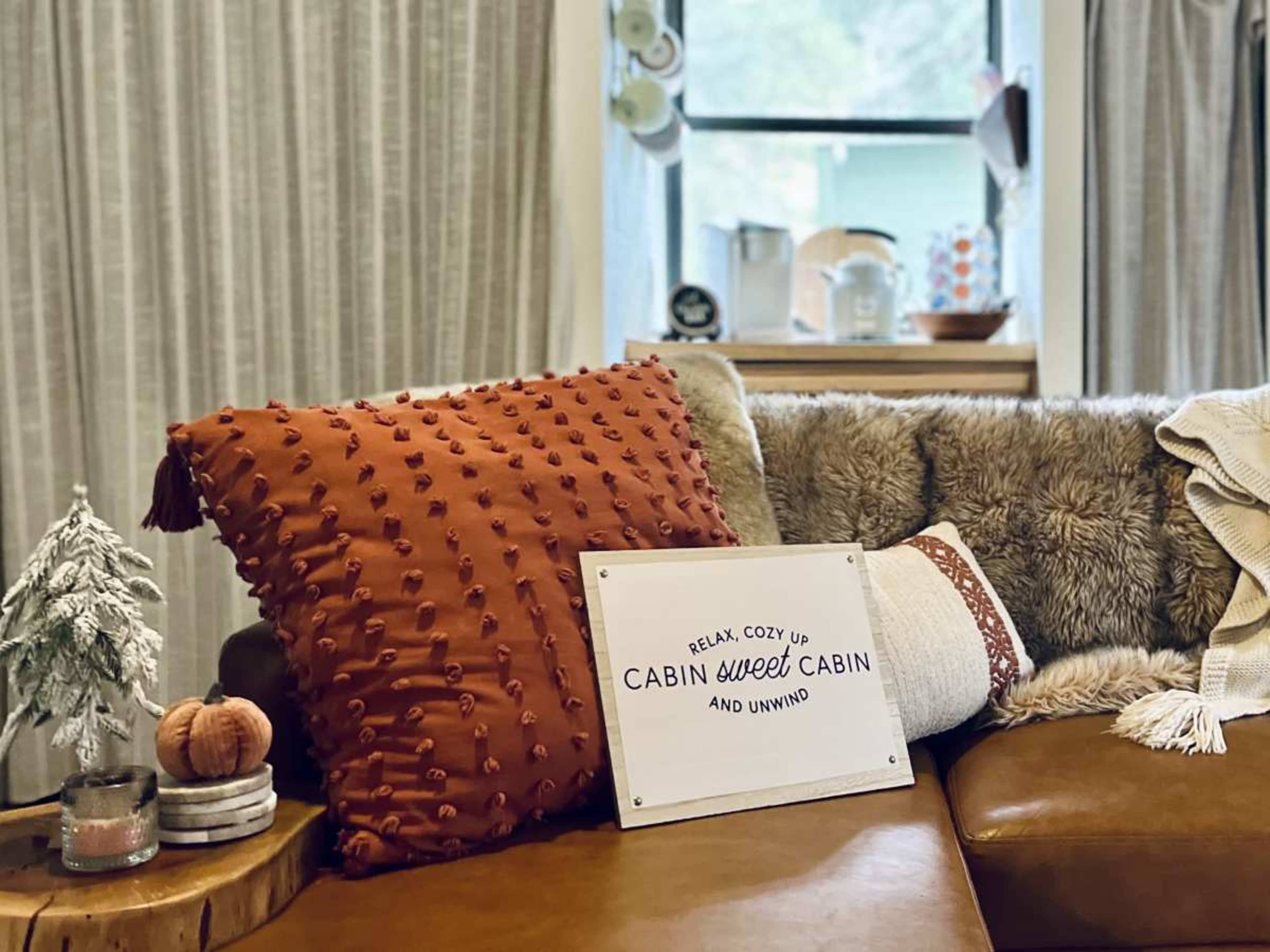 A cozy living room scene features a brown leather couch adorned with textured pillows and a decorative sign reading "Relax, Cozy Up, Cabin Sweet Cabin and Unwind."
