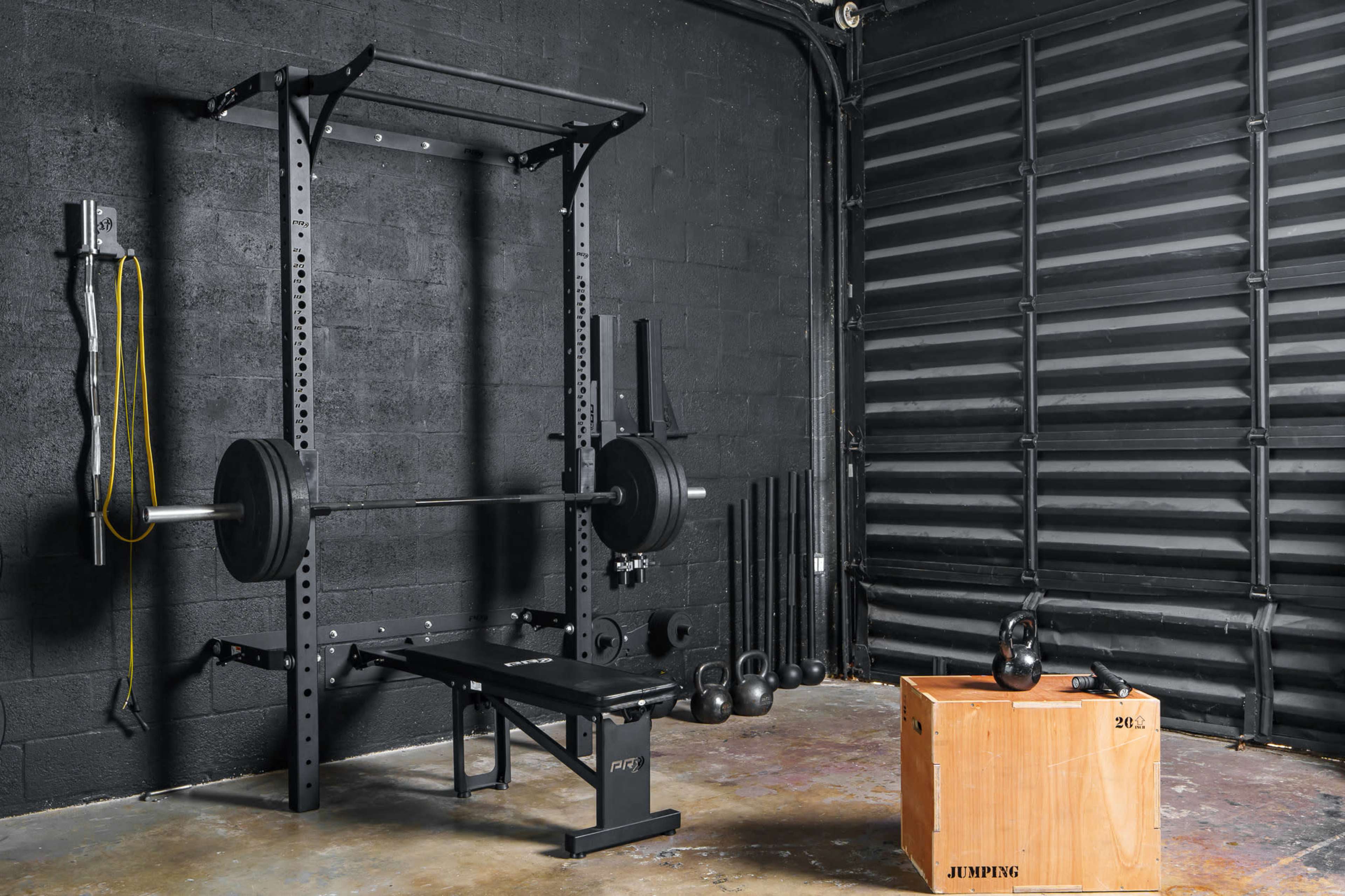 The image shows a weightlifting setup in a gym, featuring a power rack, a barbell on a bench, weights, and a wooden plyometric box.