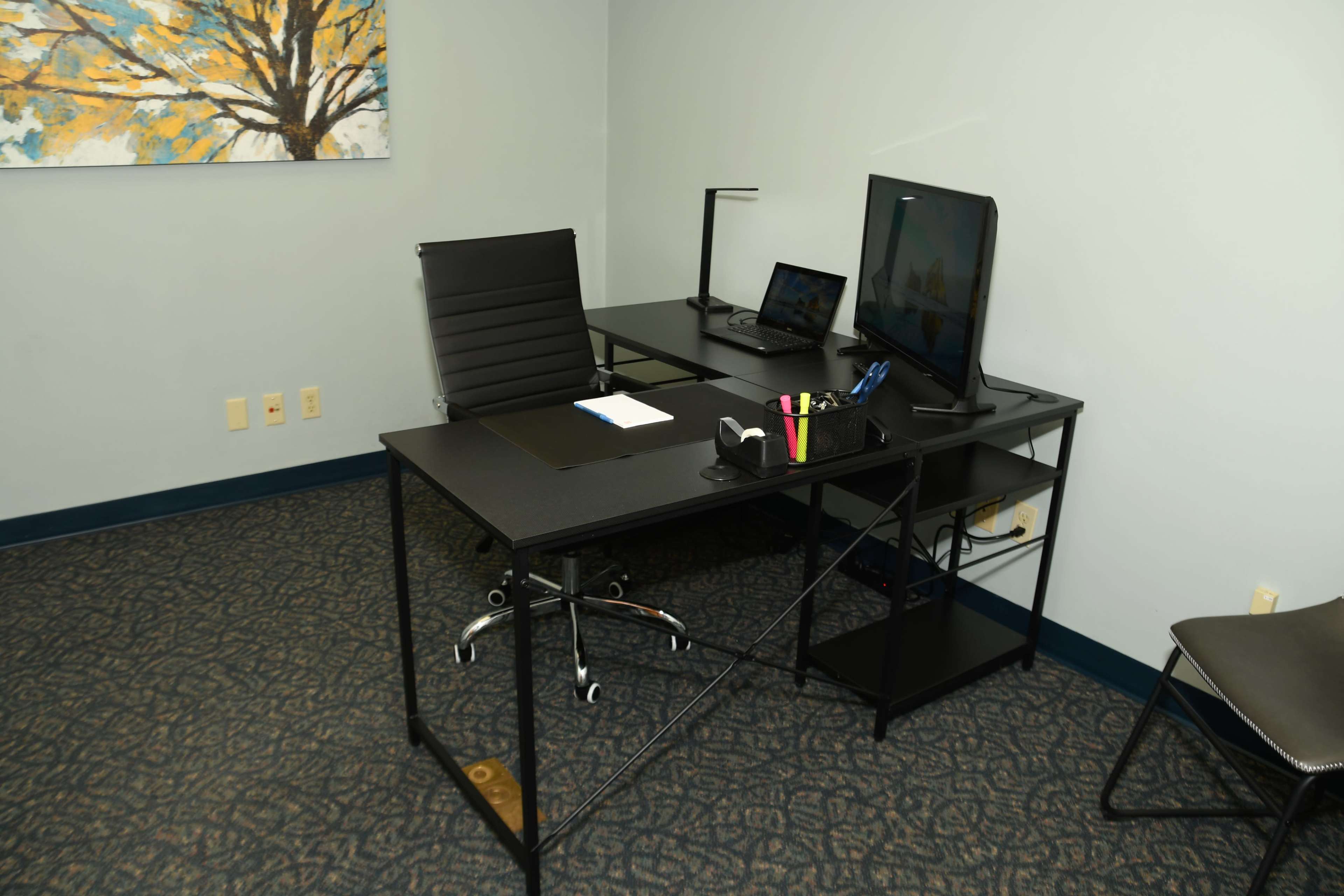 The image shows a modern office space with a black desk, an ergonomic chair, a computer monitor, a laptop, and organized office supplies on a shelf against a light blue wall.