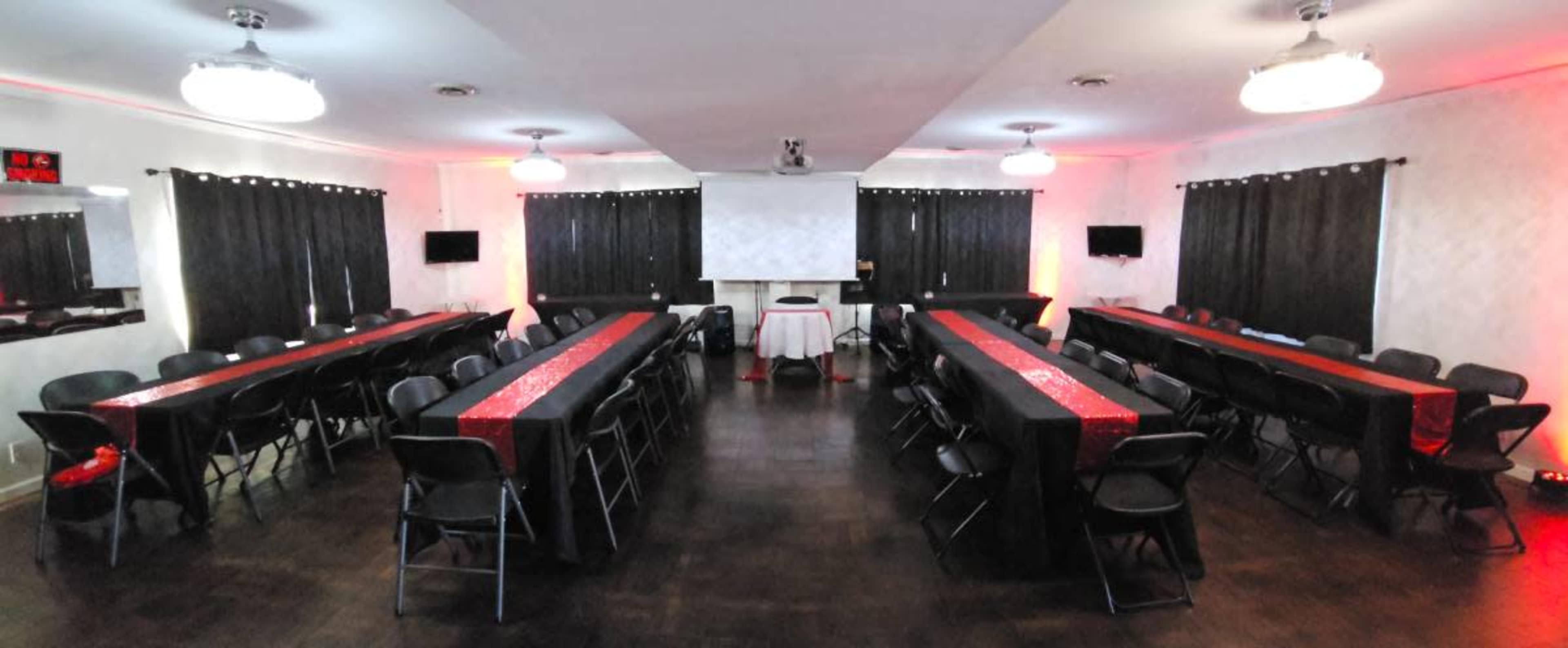 The image shows a banquet hall set up with several long tables arranged in an open space, with black chairs and decorative red accents.