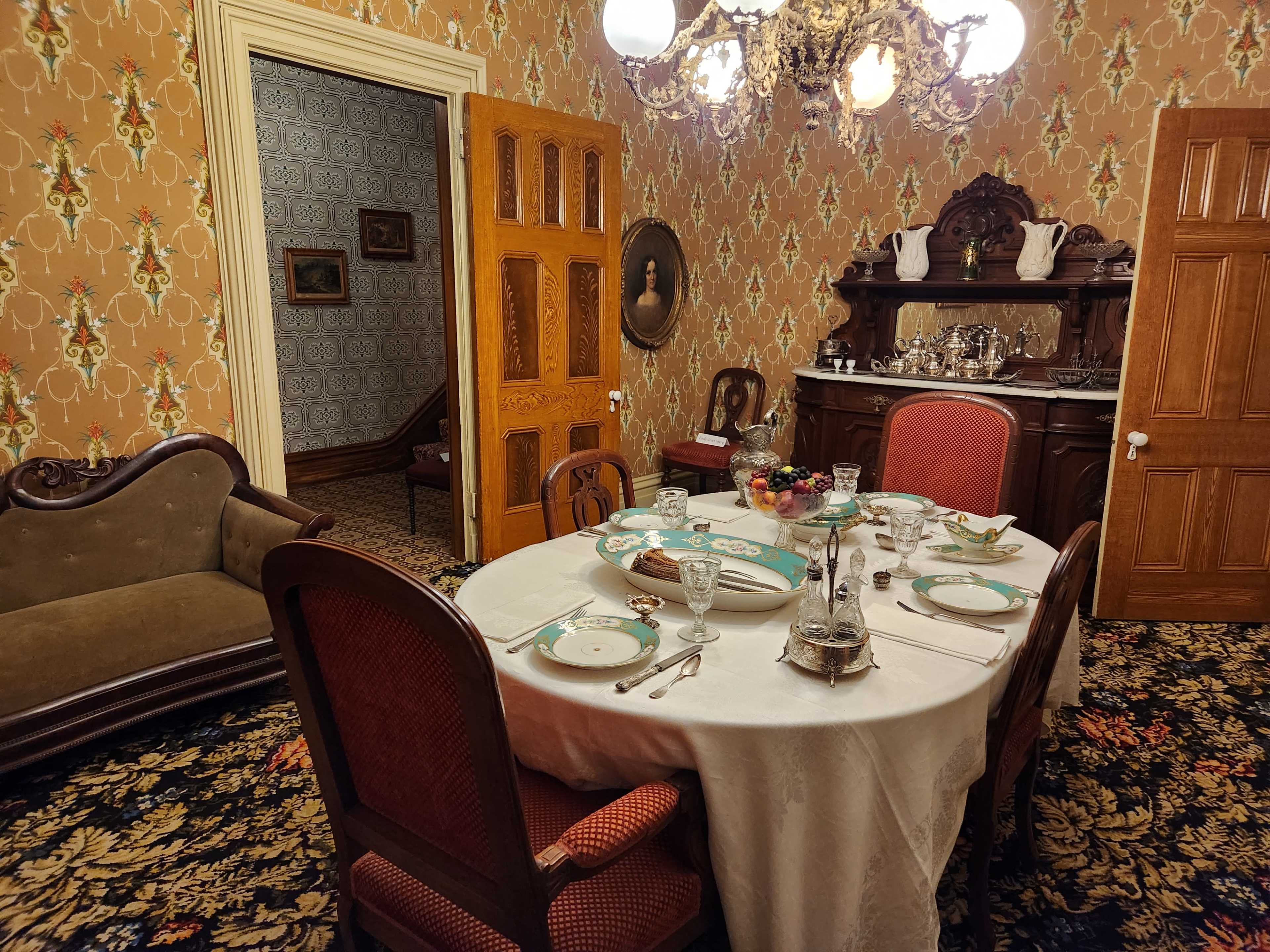 A formal dining room set with a table featuring an ornate tablecloth, china, and glassware, surrounded by wooden chairs against a backdrop of patterned wallpaper and vintage decor.