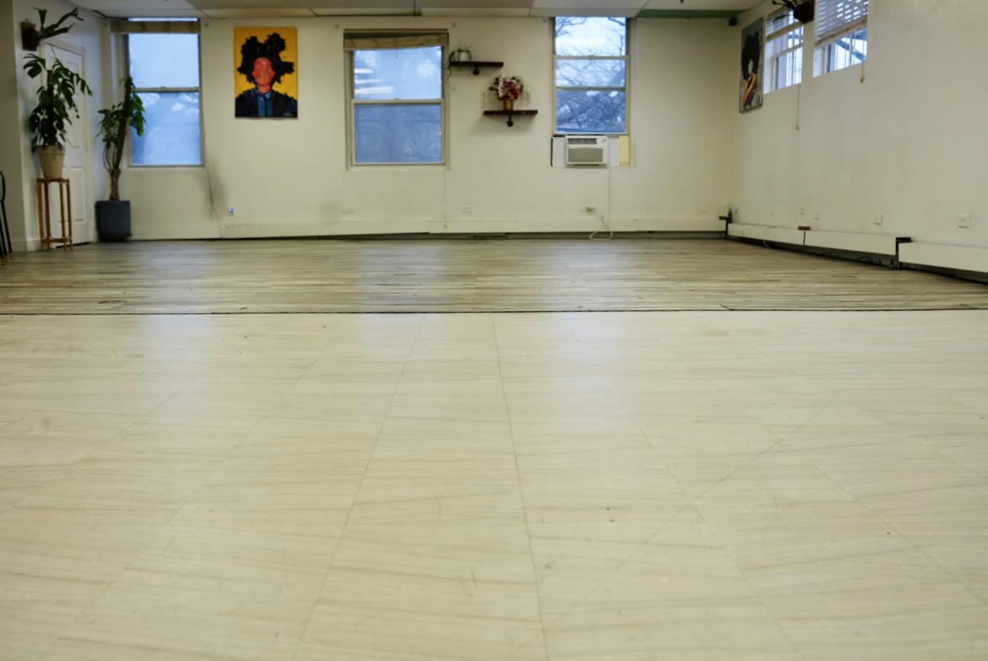 An empty indoor space with wooden flooring, large windows, and two wall-mounted portraits.