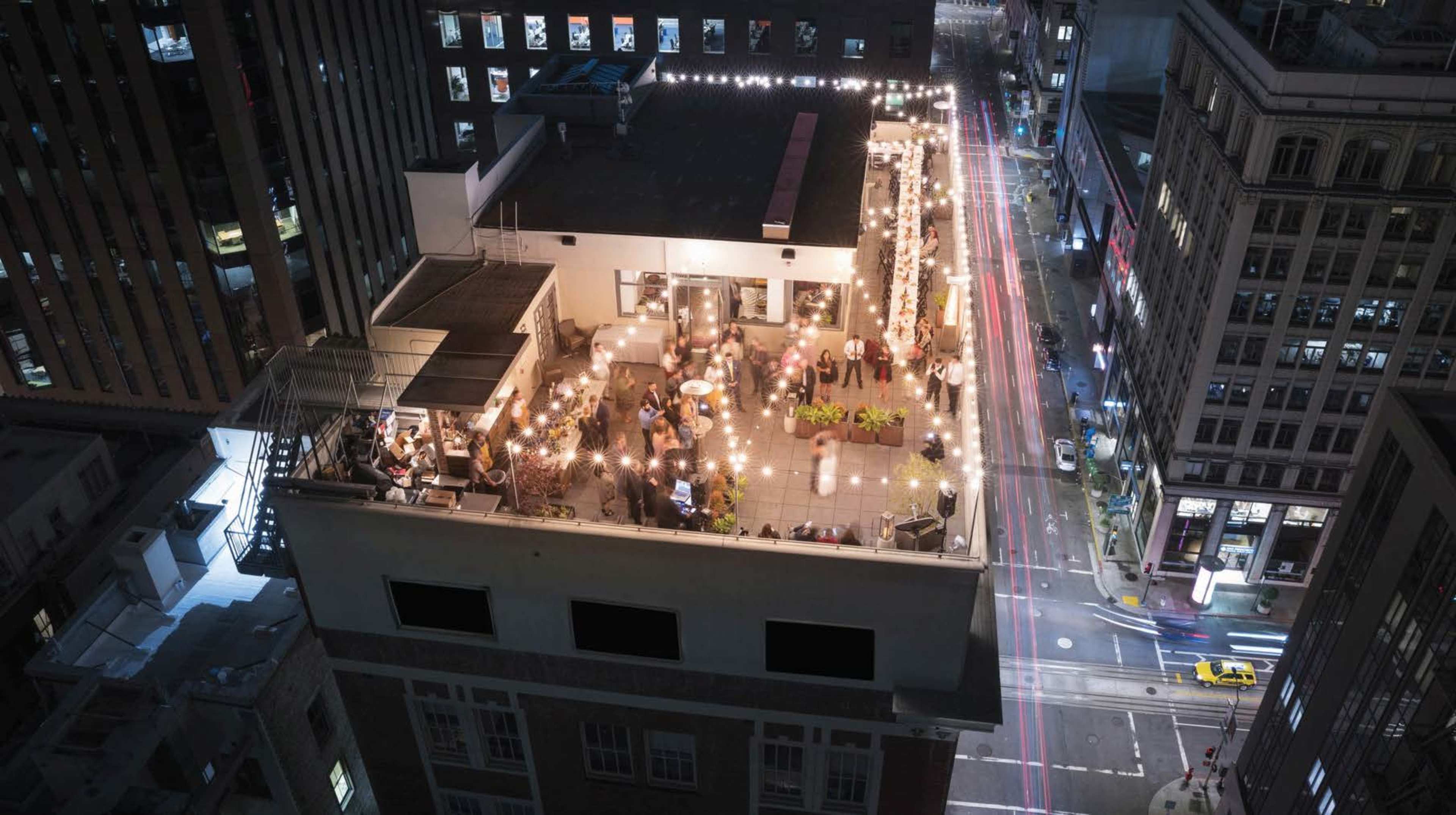 A rooftop bar filled with patrons and string lights overlooks a busy city street at night.