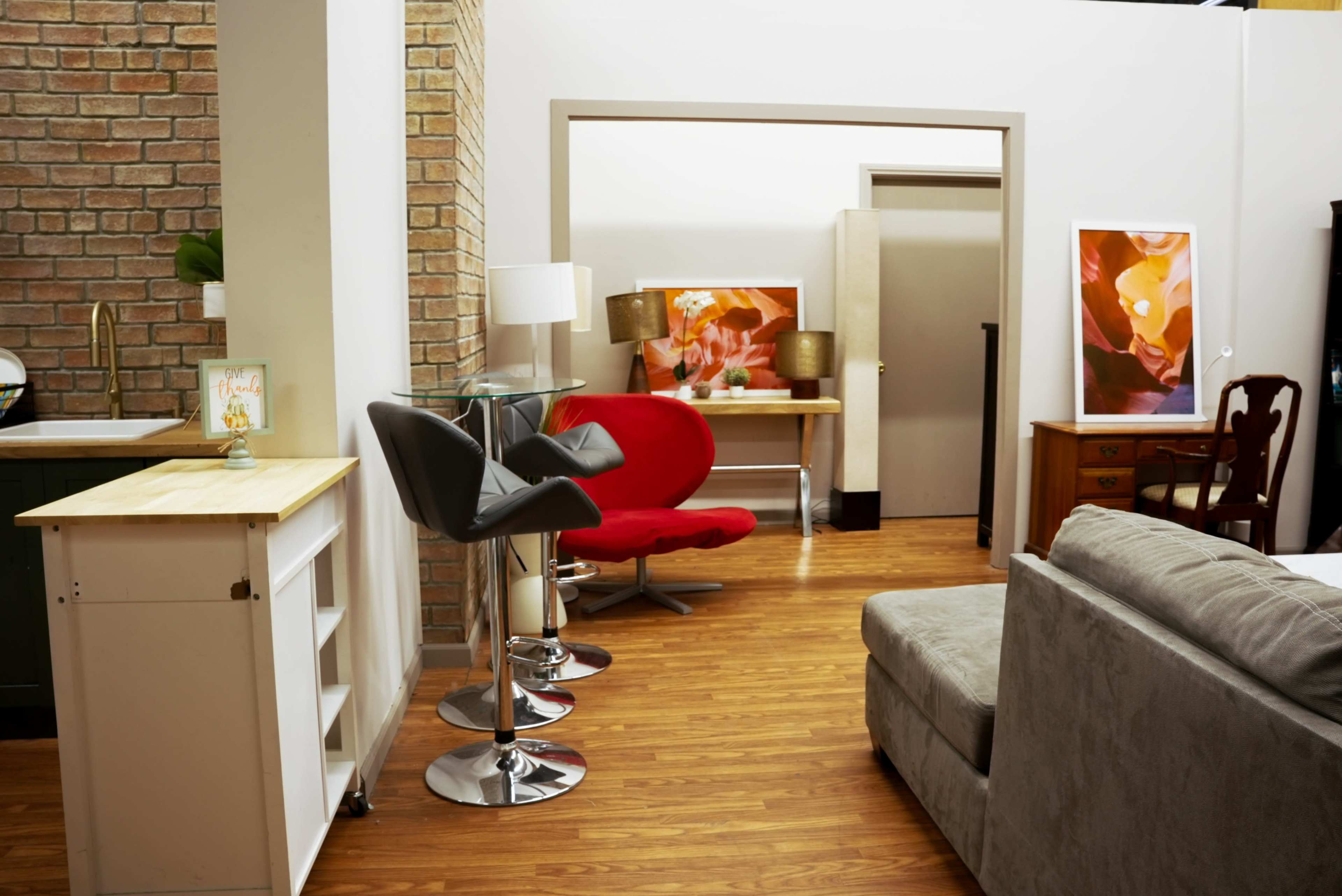 The image shows a modern interior living space featuring a gray sofa, a red chair, a bar table with high stools, and artwork on the walls.