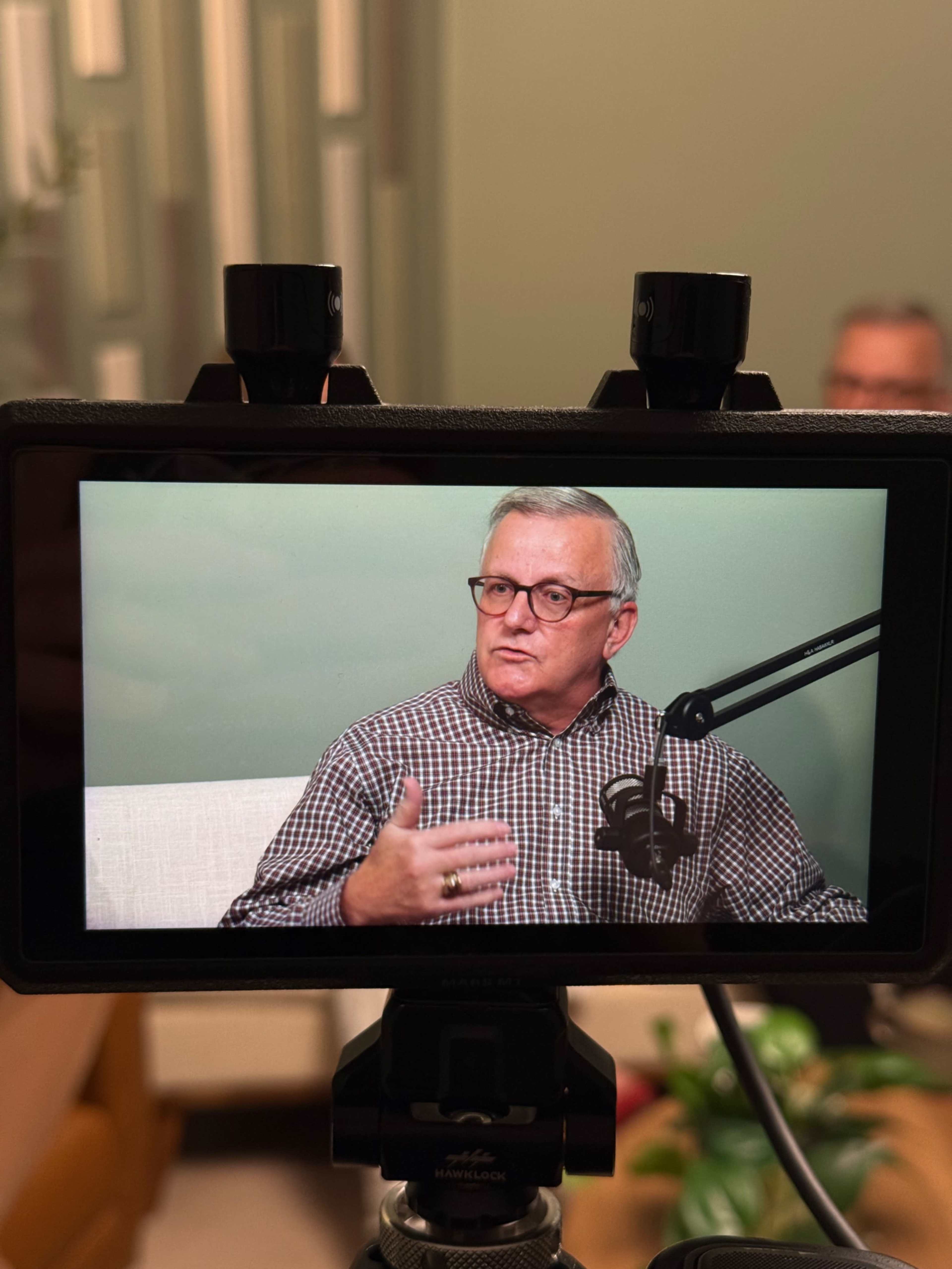 A man in a checkered shirt is speaking while sitting in front of a microphone, captured through a camera screen.