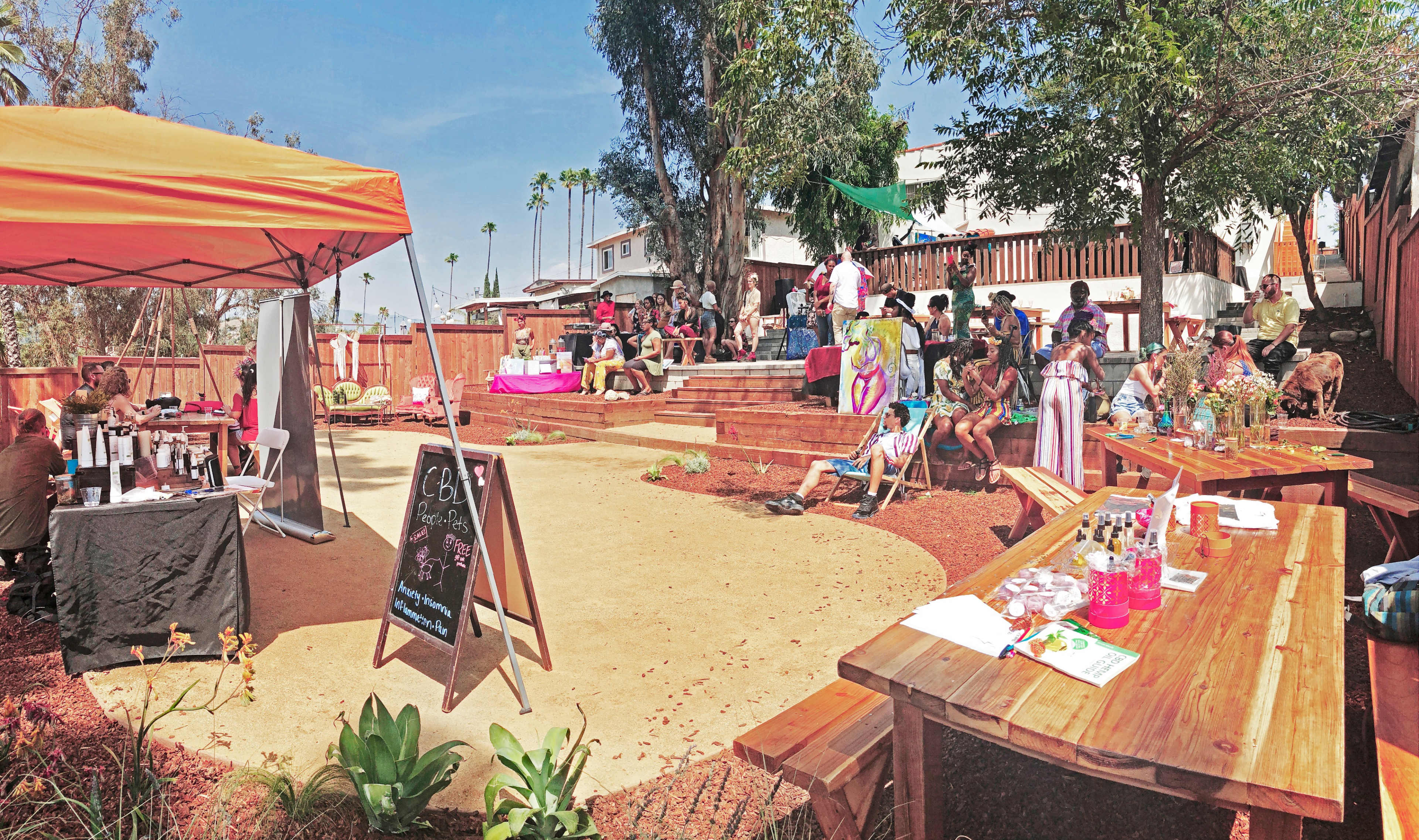 A lively outdoor gathering takes place in a spacious backyard, featuring wooden seating areas, a sandy centerpiece, and a tented booth serving beverages.
