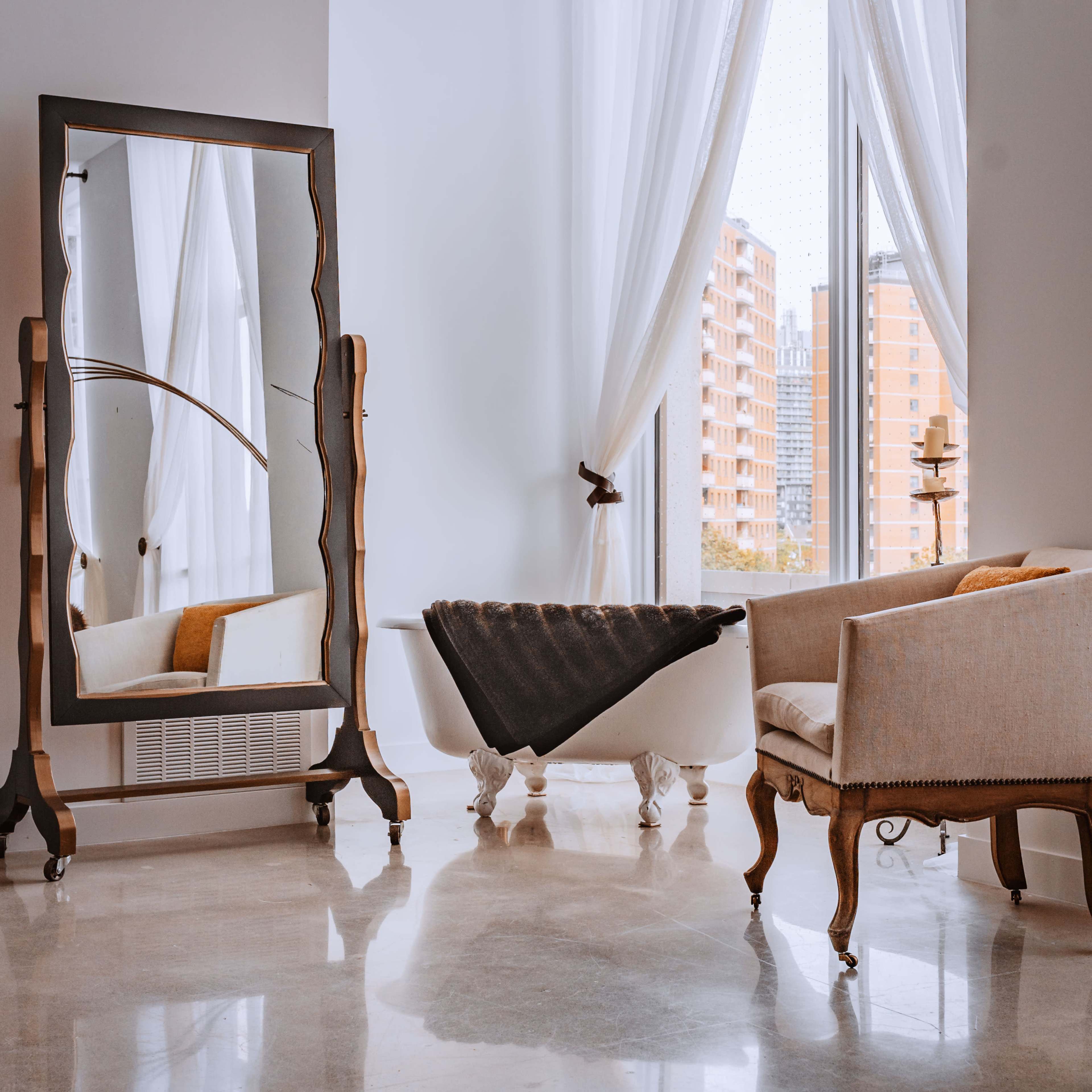 The image shows a bright room with large windows, featuring a full-length mirror, a vintage armchair, and a freestanding bathtub decorated with a gray blanket.