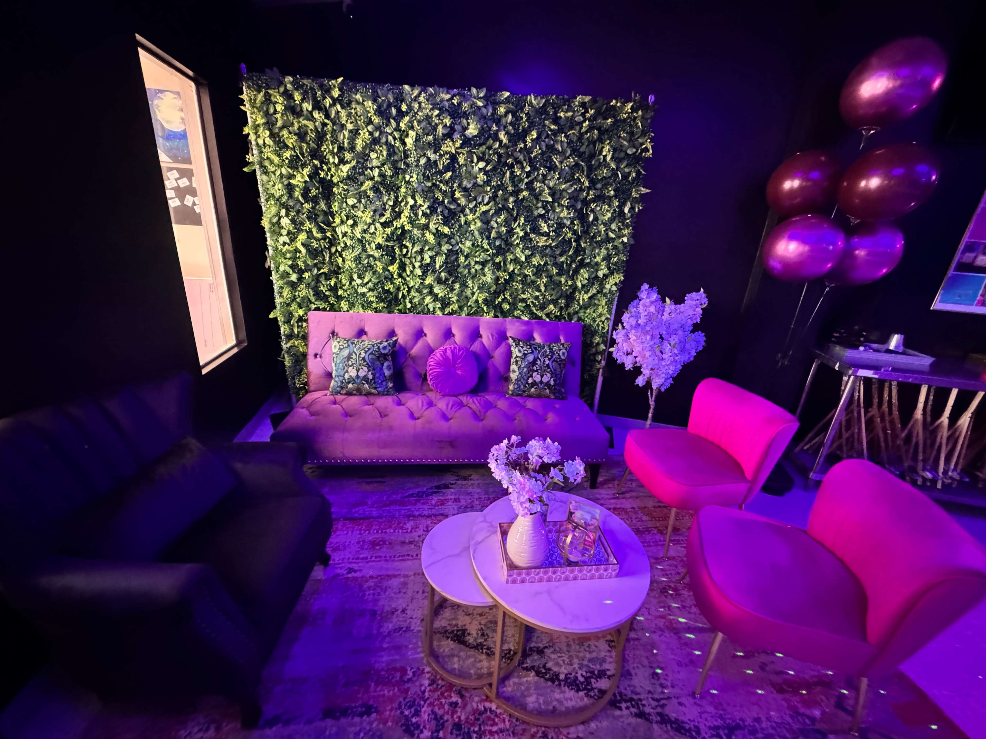 The image shows a vibrant seating area featuring a purple sofa, pink chairs, and a decorative green wall, with balloons and flowers enhancing the decor.
