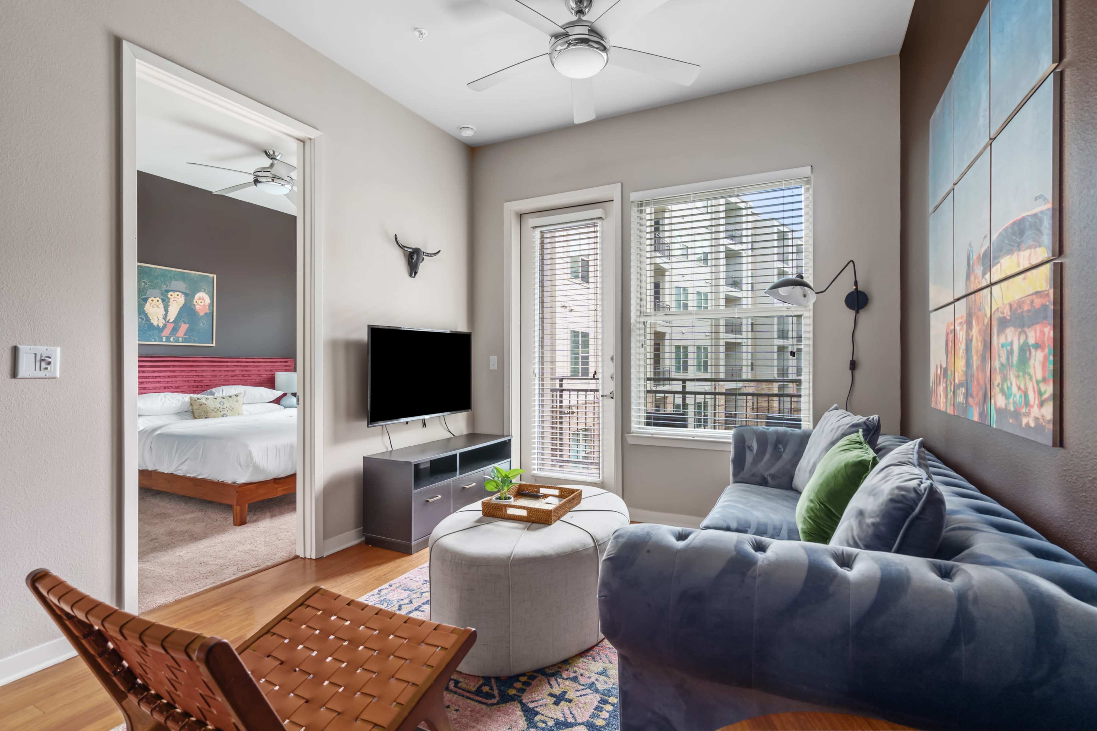 A living room features a blue sofa, a round ottoman, a television on a stand, and an adjoining bedroom visible through a set of double doors.