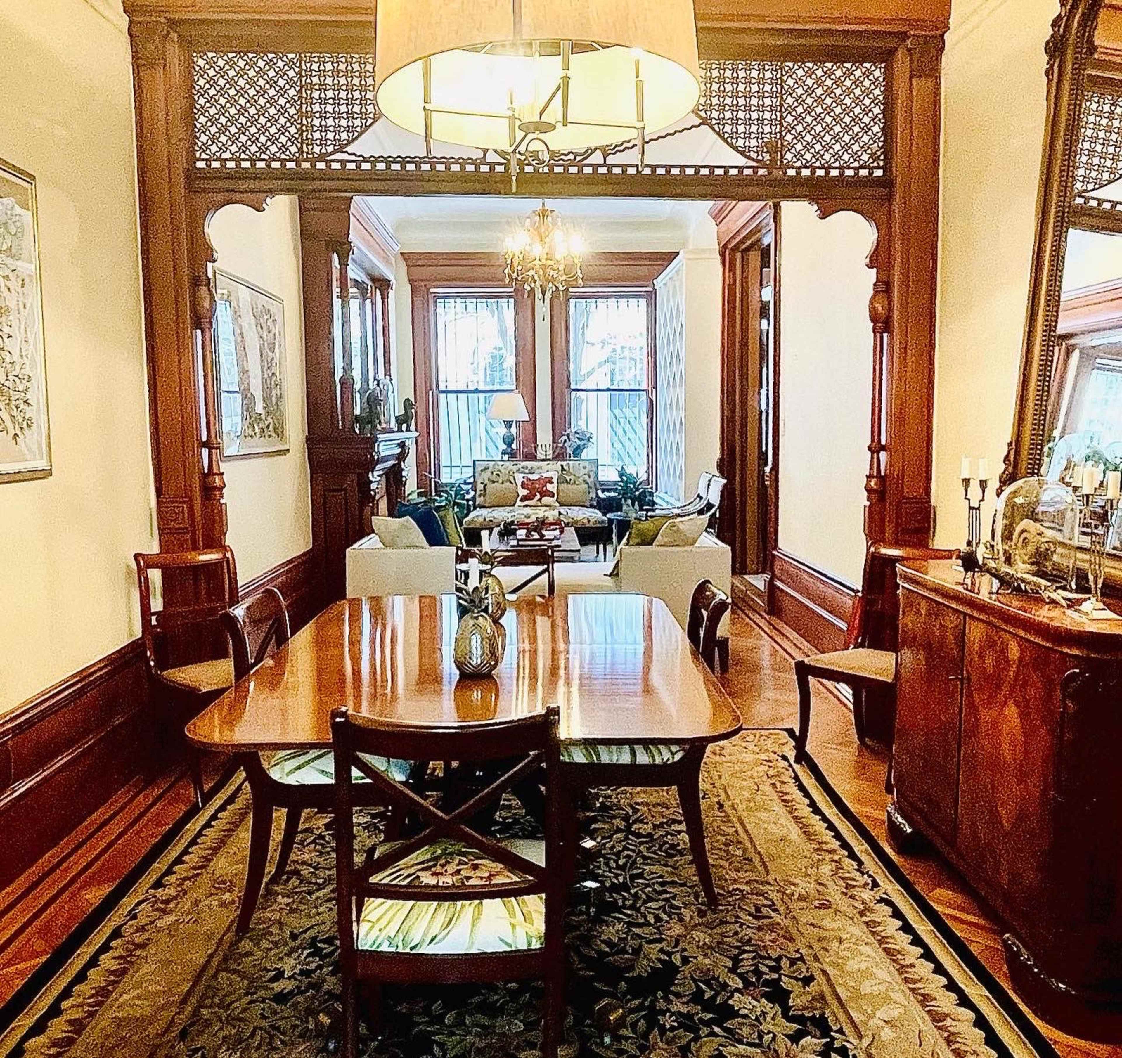 A wood-paneled dining room with a table set for six, leading into a brightly lit living area furnished with sofas and decorative plants.