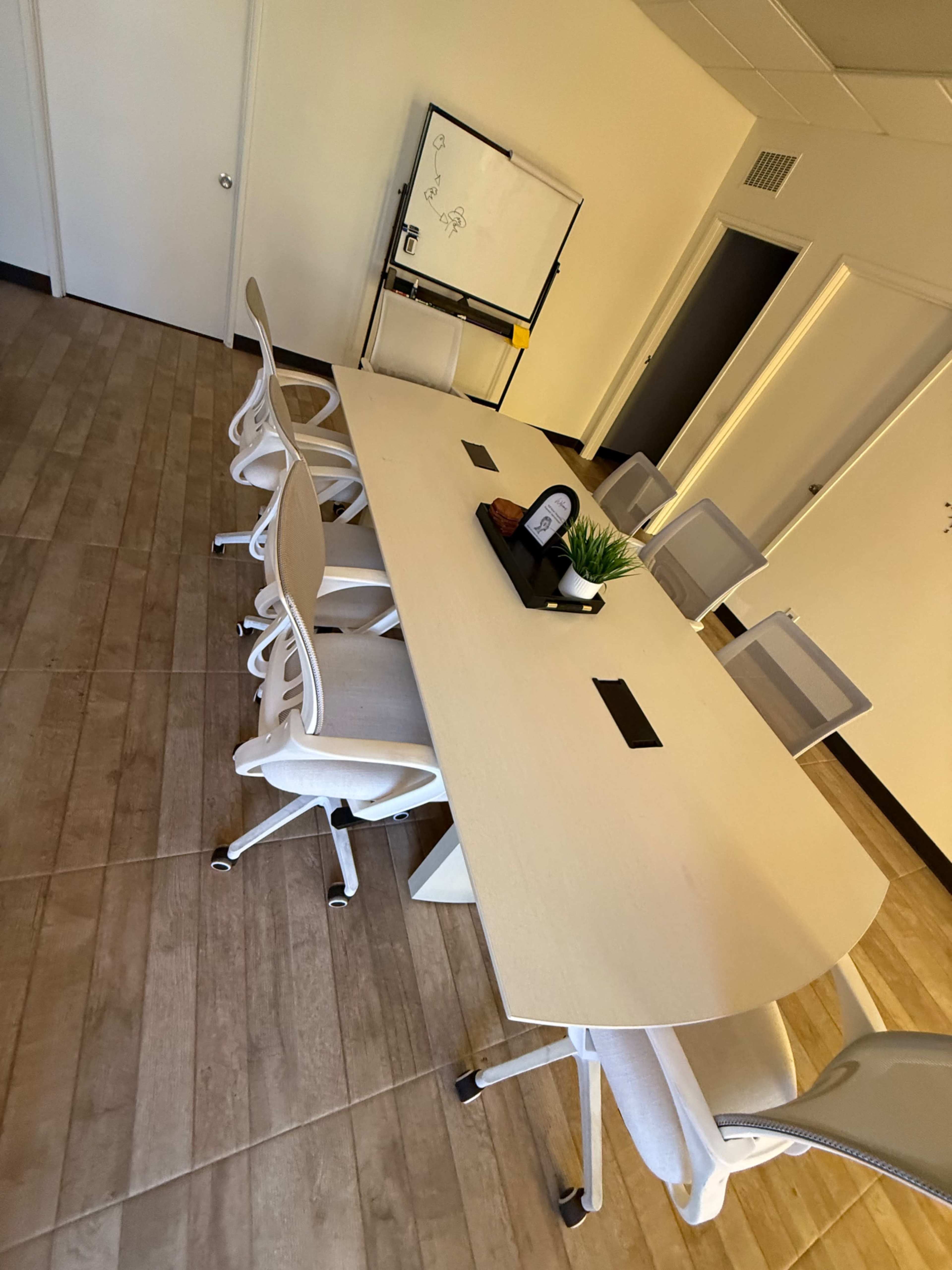The image shows a modern conference room with a long wooden table, several office chairs, and a whiteboard on the wall.