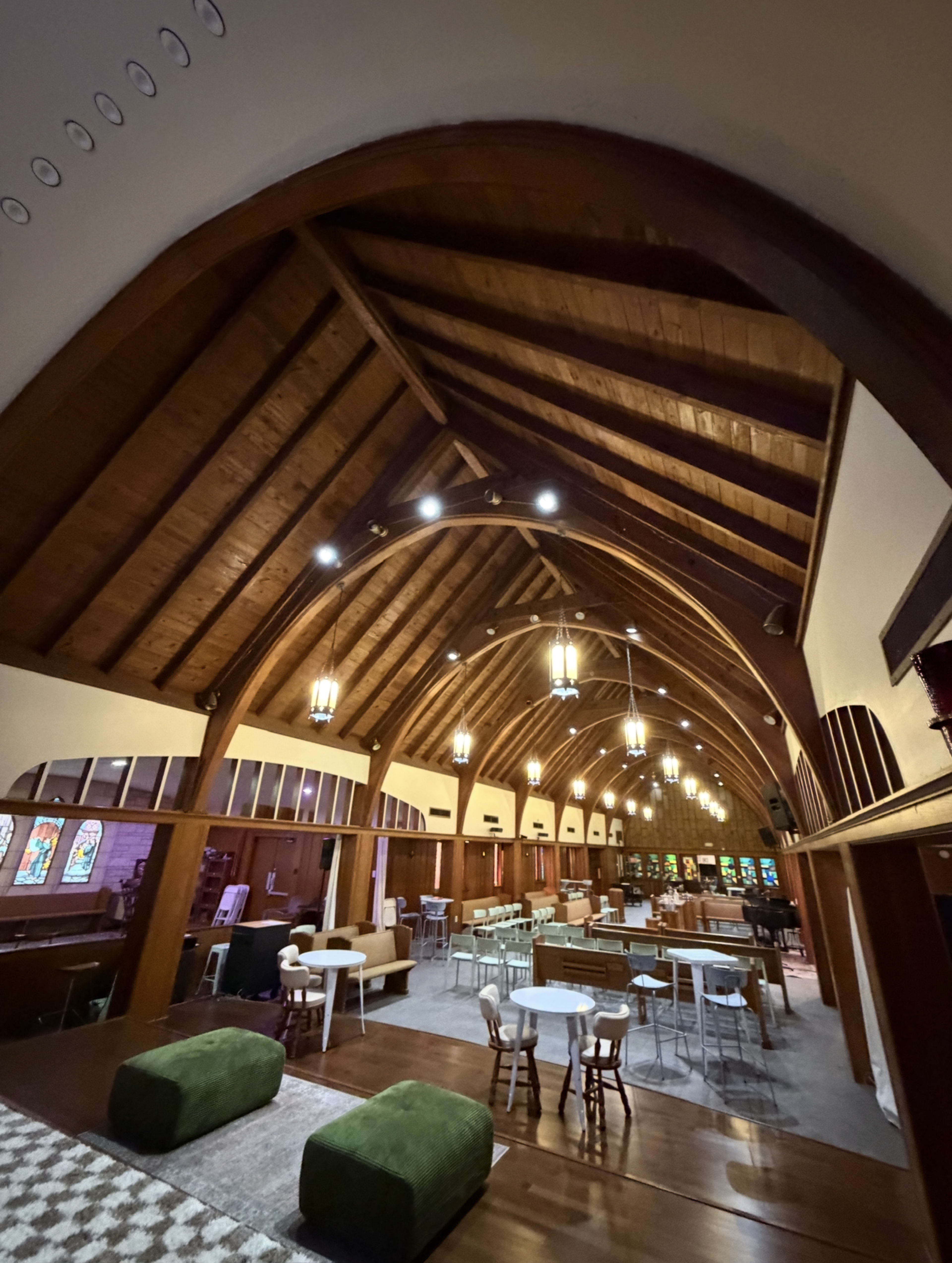The interior of a church features a high wooden ceiling, stained glass windows, and a mix of seating arrangements including tables and chairs.