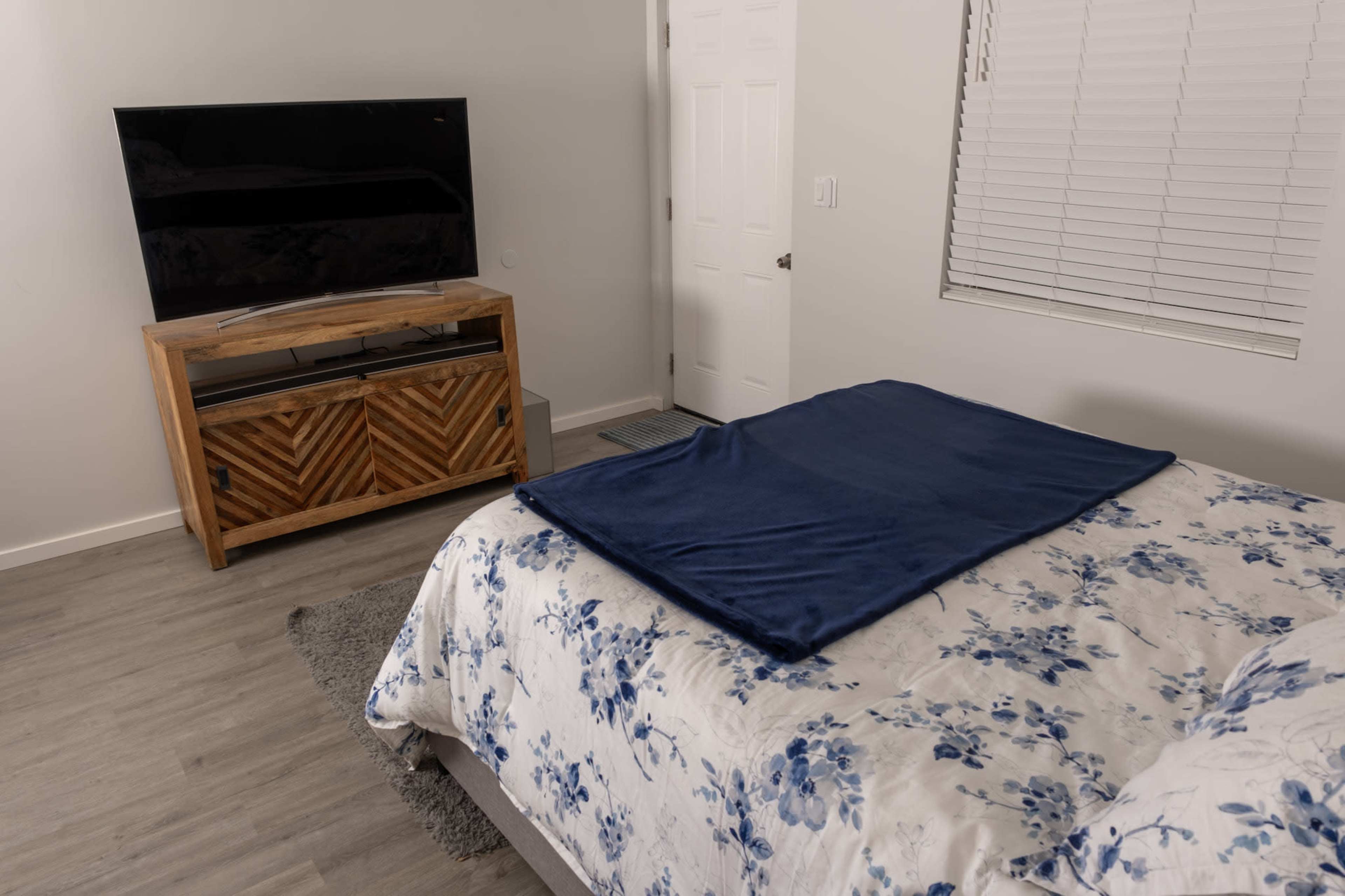 The image shows a bedroom with a bed covered in a floral-patterned blanket, a wooden cabinet beneath a television, and a closed door leading to another room.