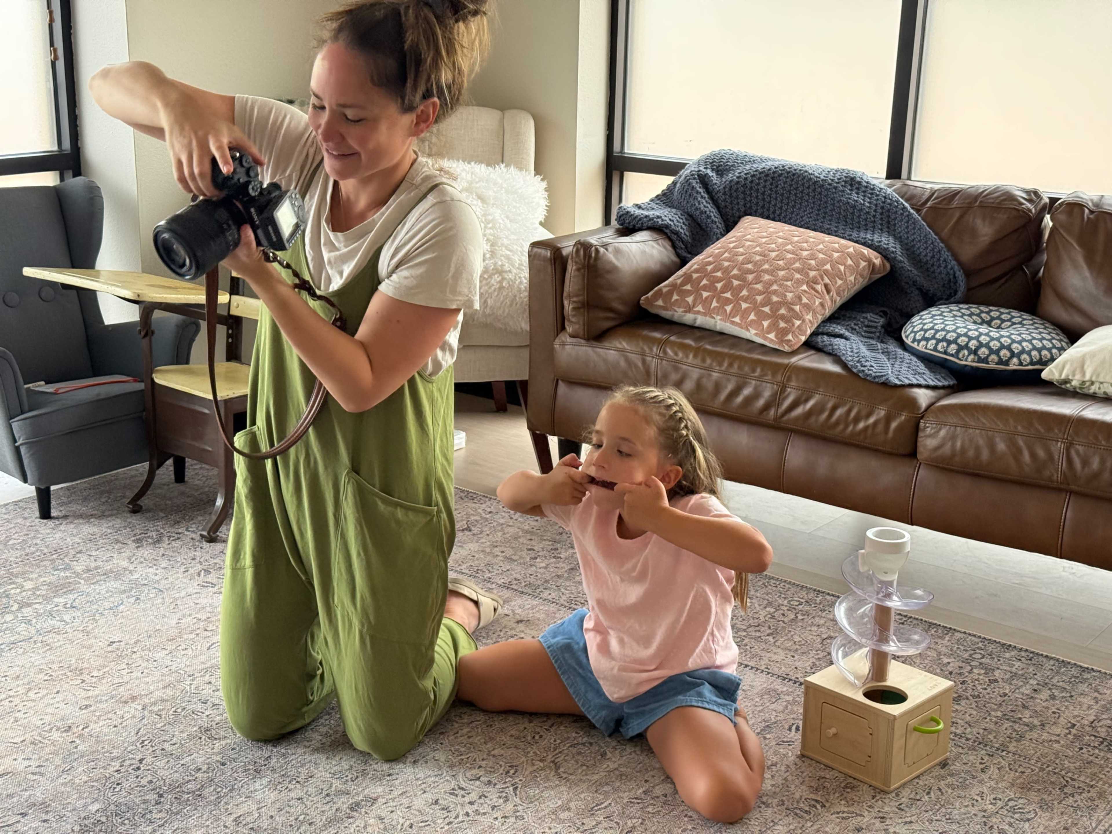 A woman takes pictures with a camera while a girl poses playfully on the floor beside her.
