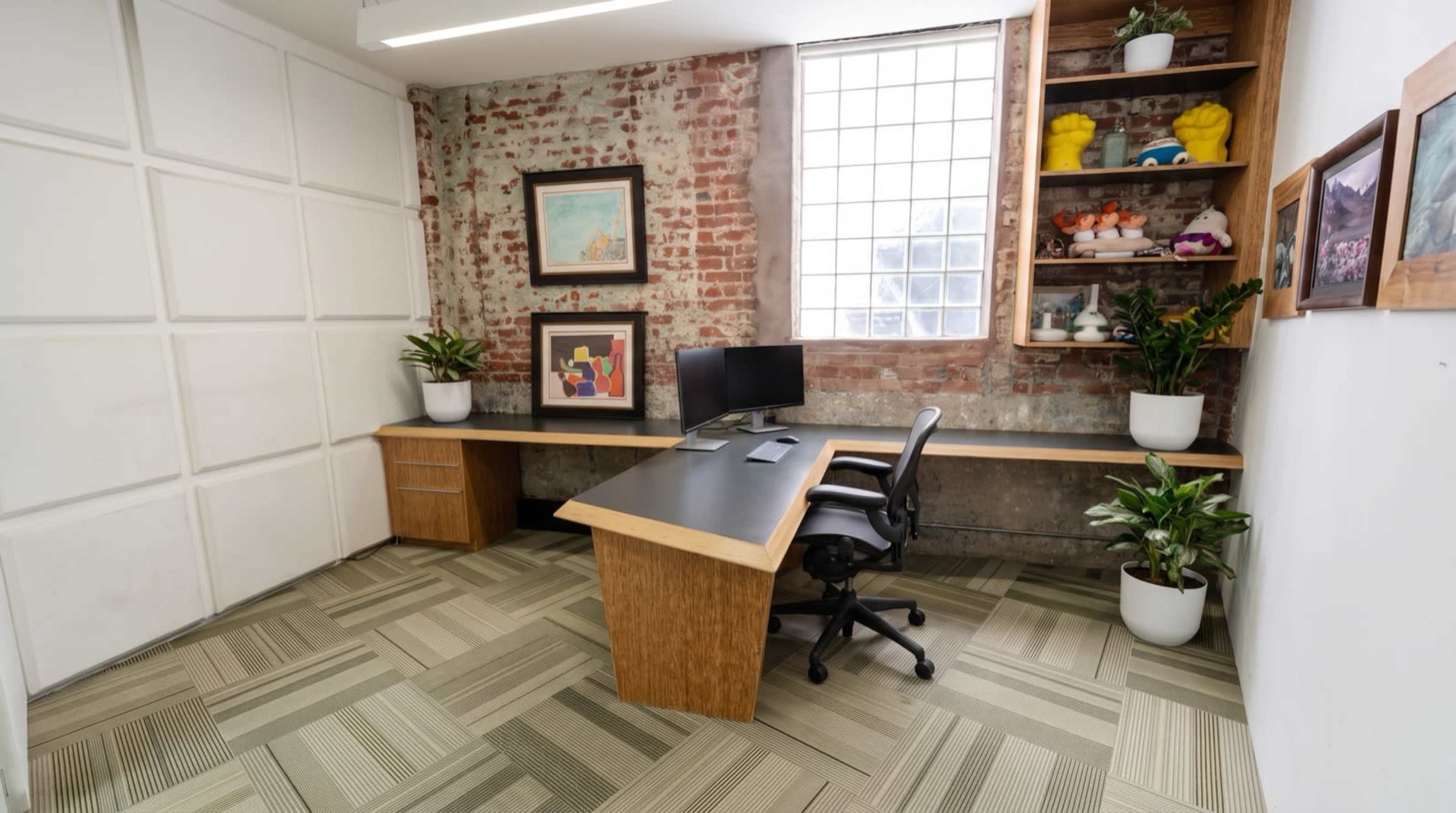 The image shows a modern office space featuring a wooden desk, a computer monitor, and several plants against a backdrop of exposed brick walls and a large window.