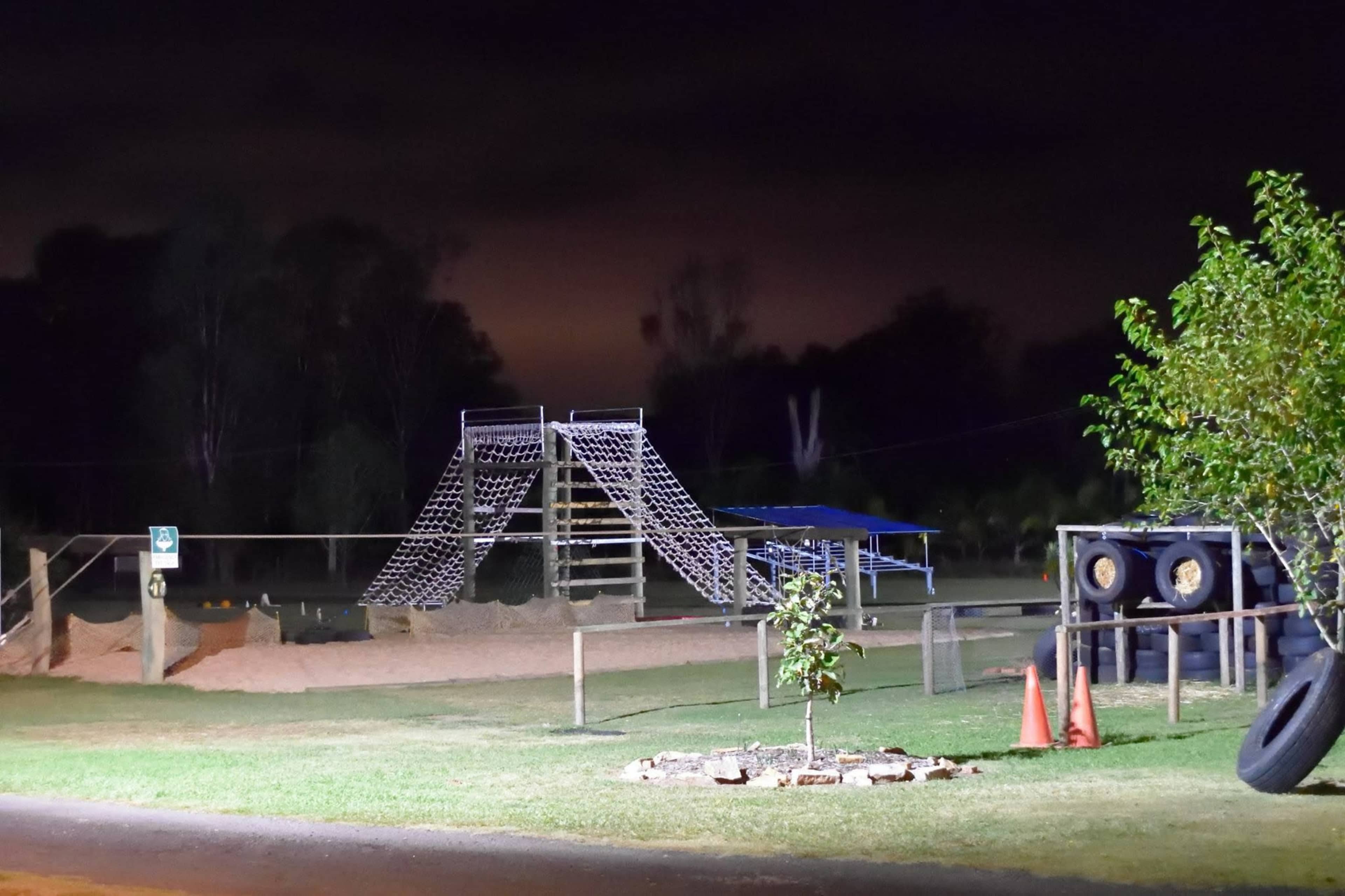 A playground at night featuring a large climbing structure, a sand area, and surrounding trees.