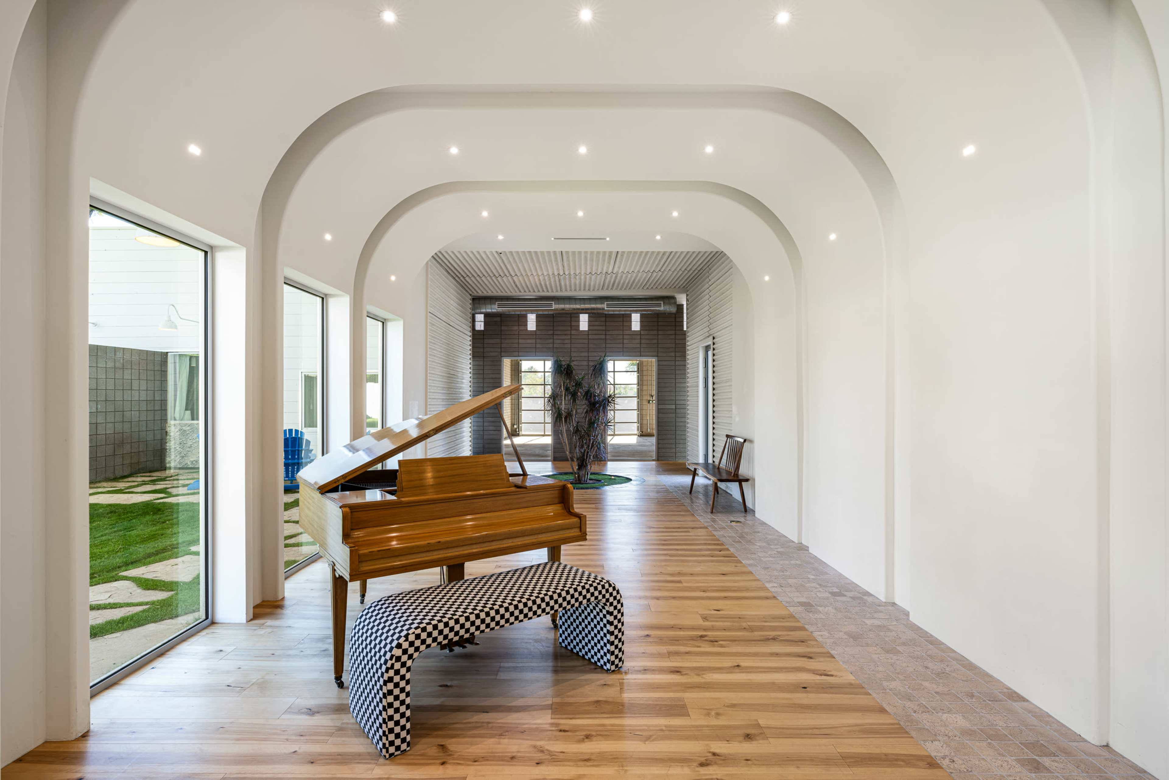 A wooden piano with a checkered bench is positioned in a spacious hallway with arched ceilings and large windows.