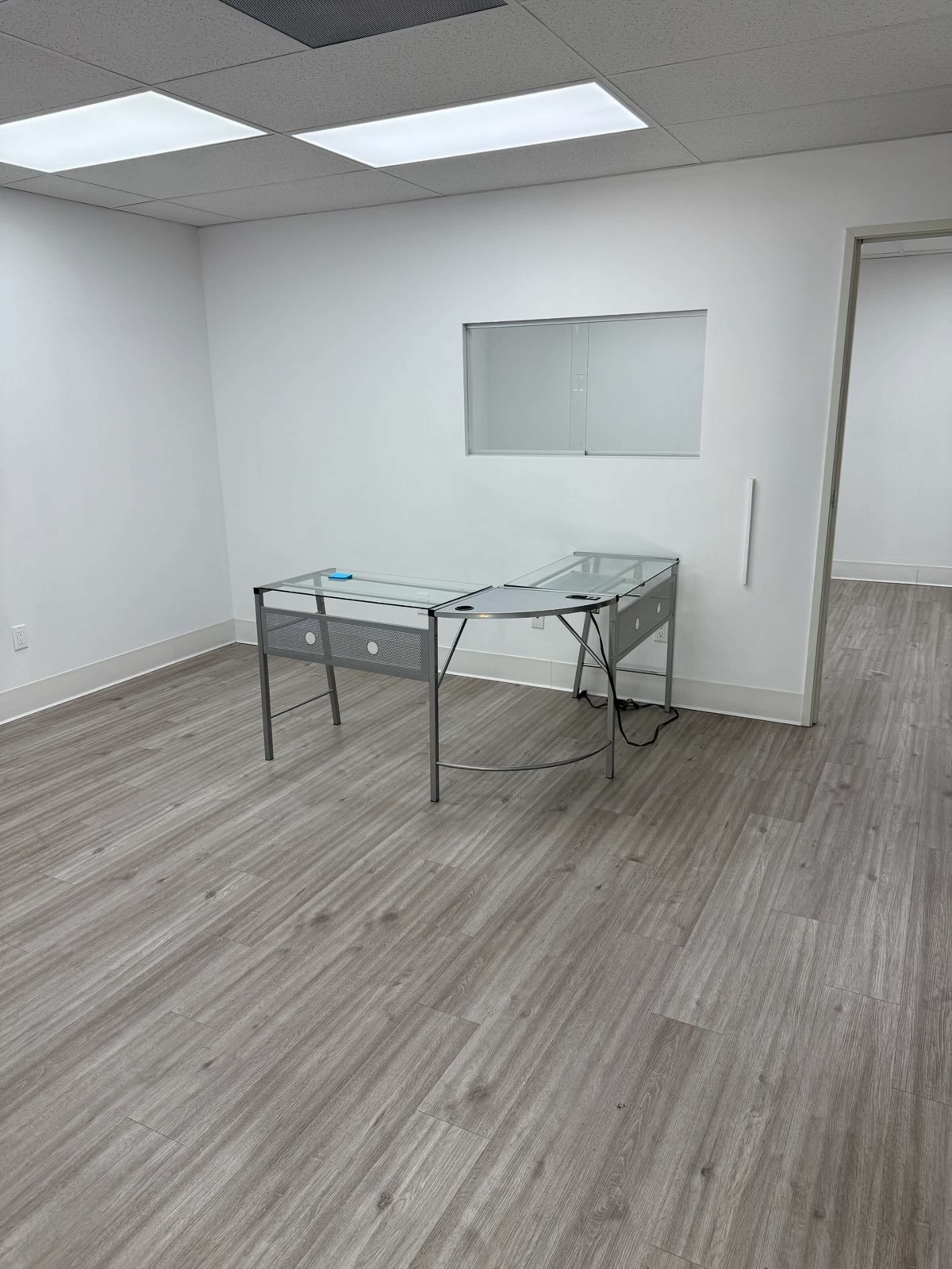 The image shows a sparse office space featuring a glass desk, a wall-mounted window, and light-colored wood flooring.