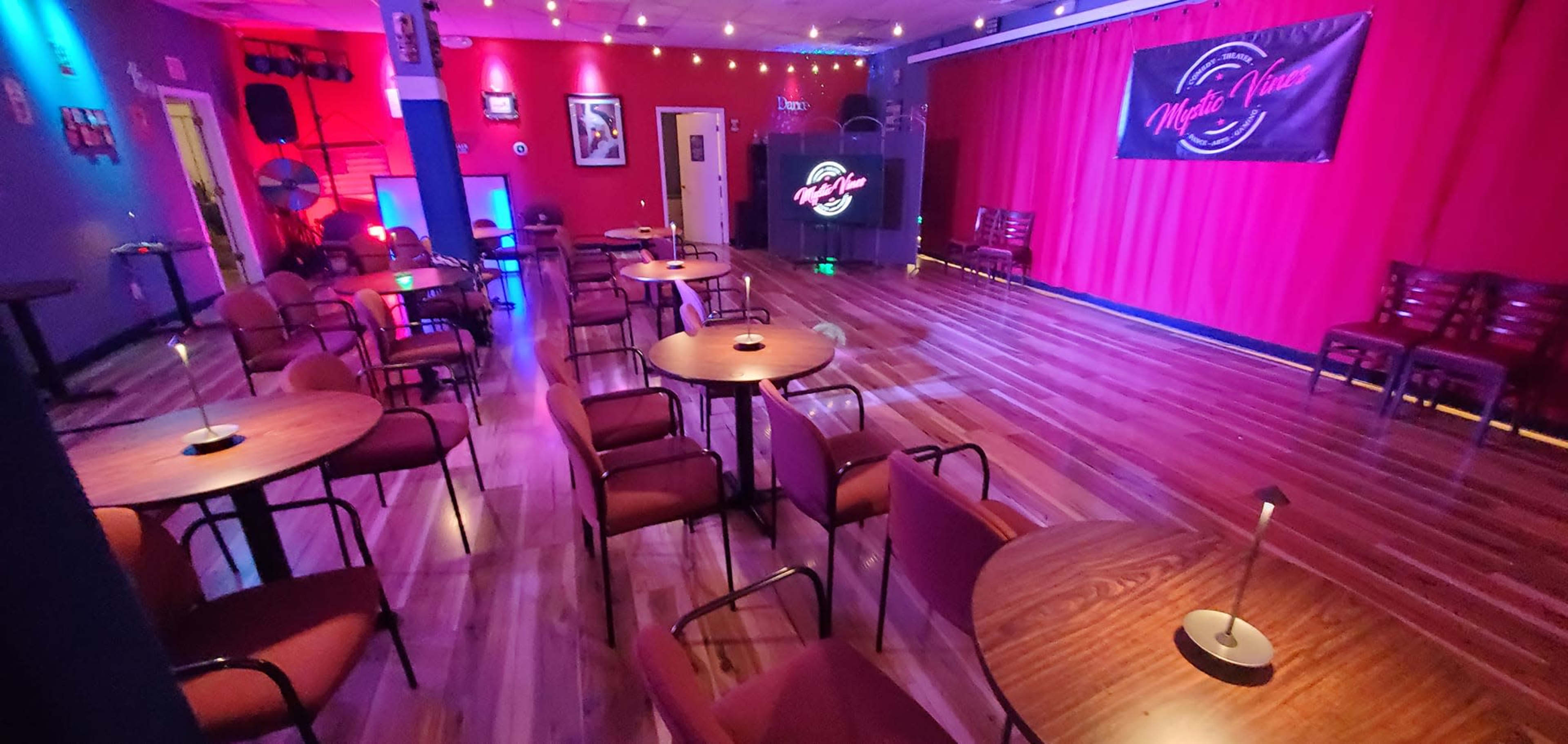 The image shows a dimly lit performance venue with several empty tables and chairs arranged in front of a small stage, adorned with red curtains and decorative lighting.